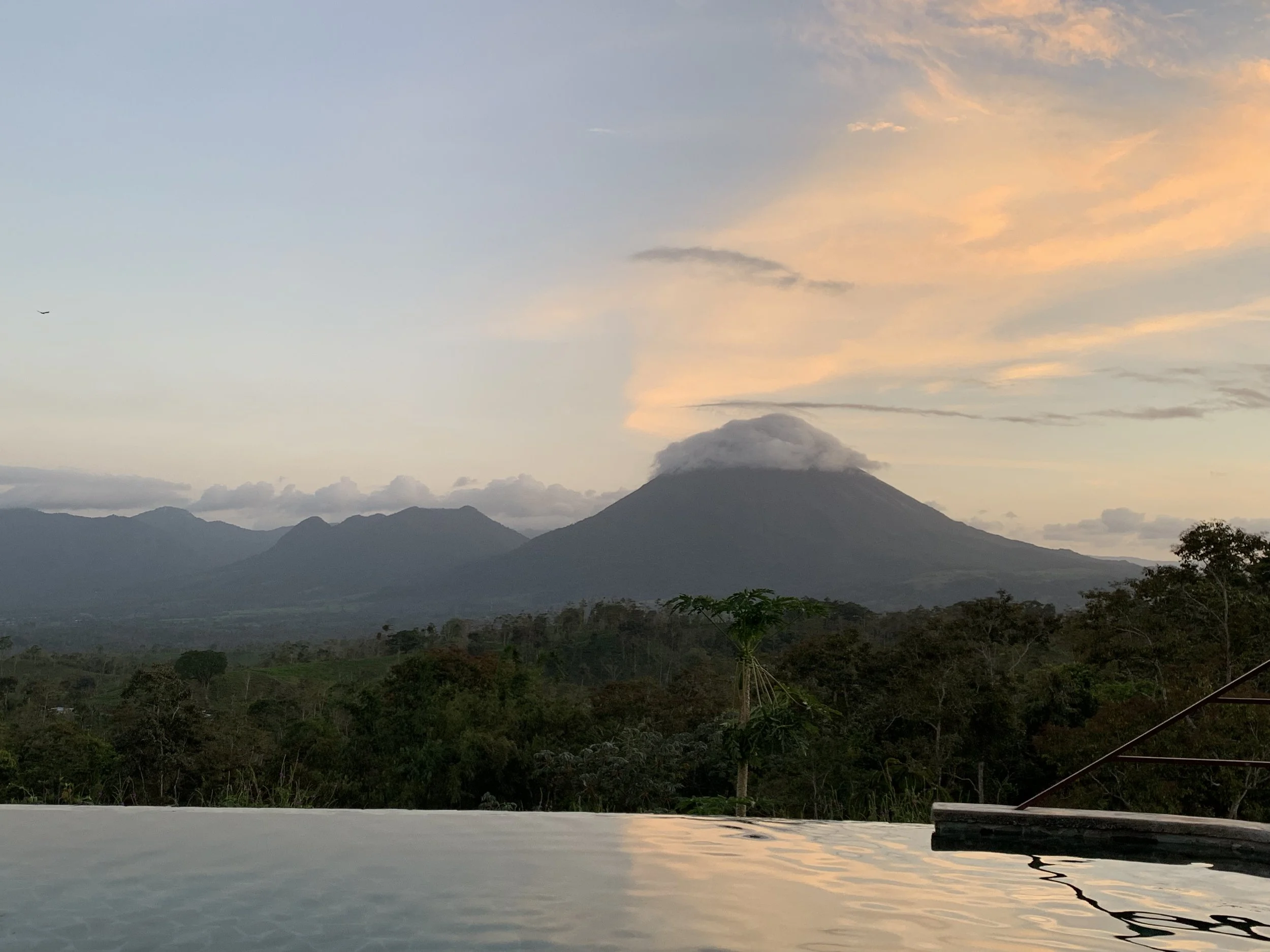 Scenic view of a mountain landscape at sunset with a smoking volcano in the background, trees in the foreground, and a reflective water surface in the lower part of the image.