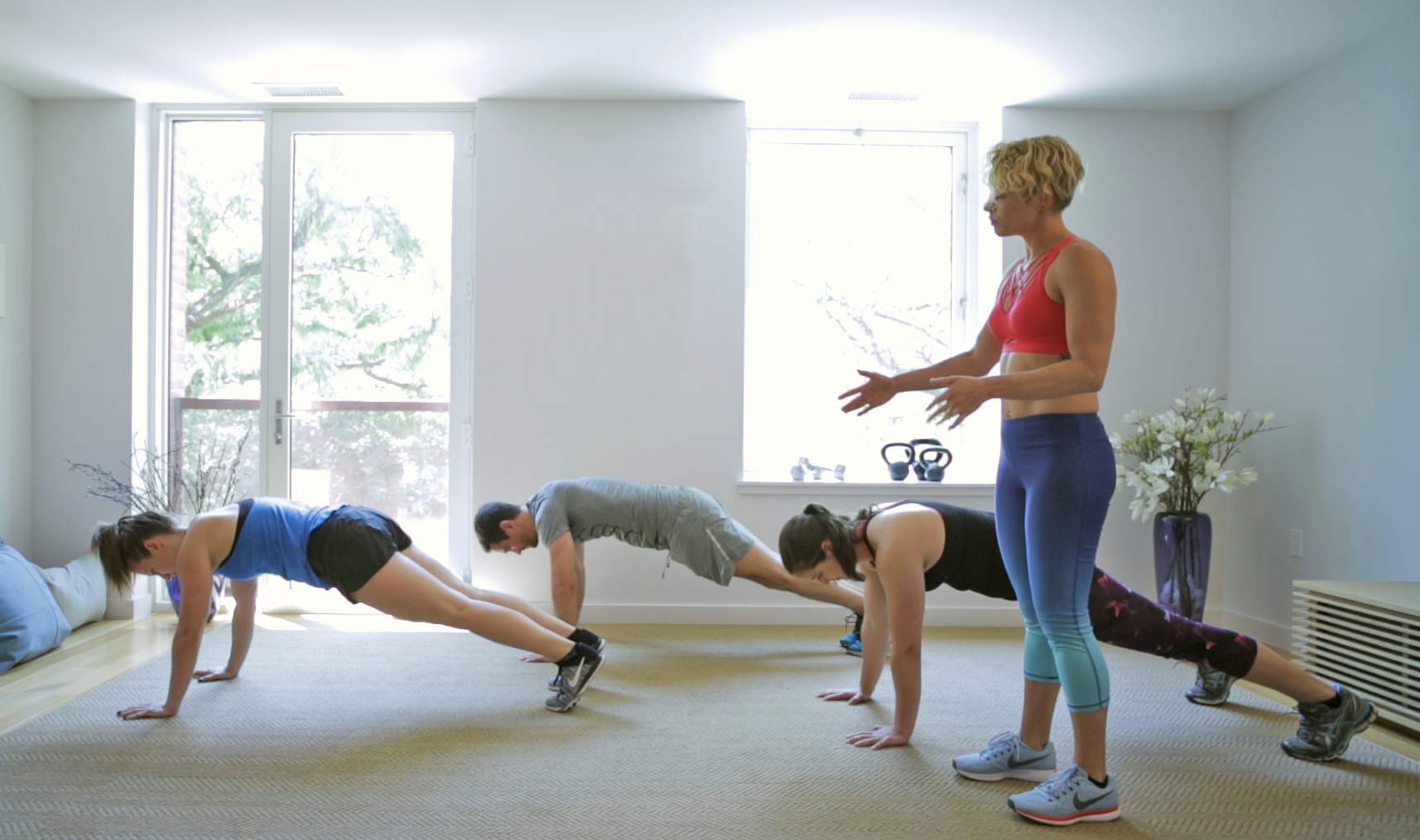 A woman leading a group workout class with four individuals doing push-ups in a bright, airy room with large windows and a door leading outside.