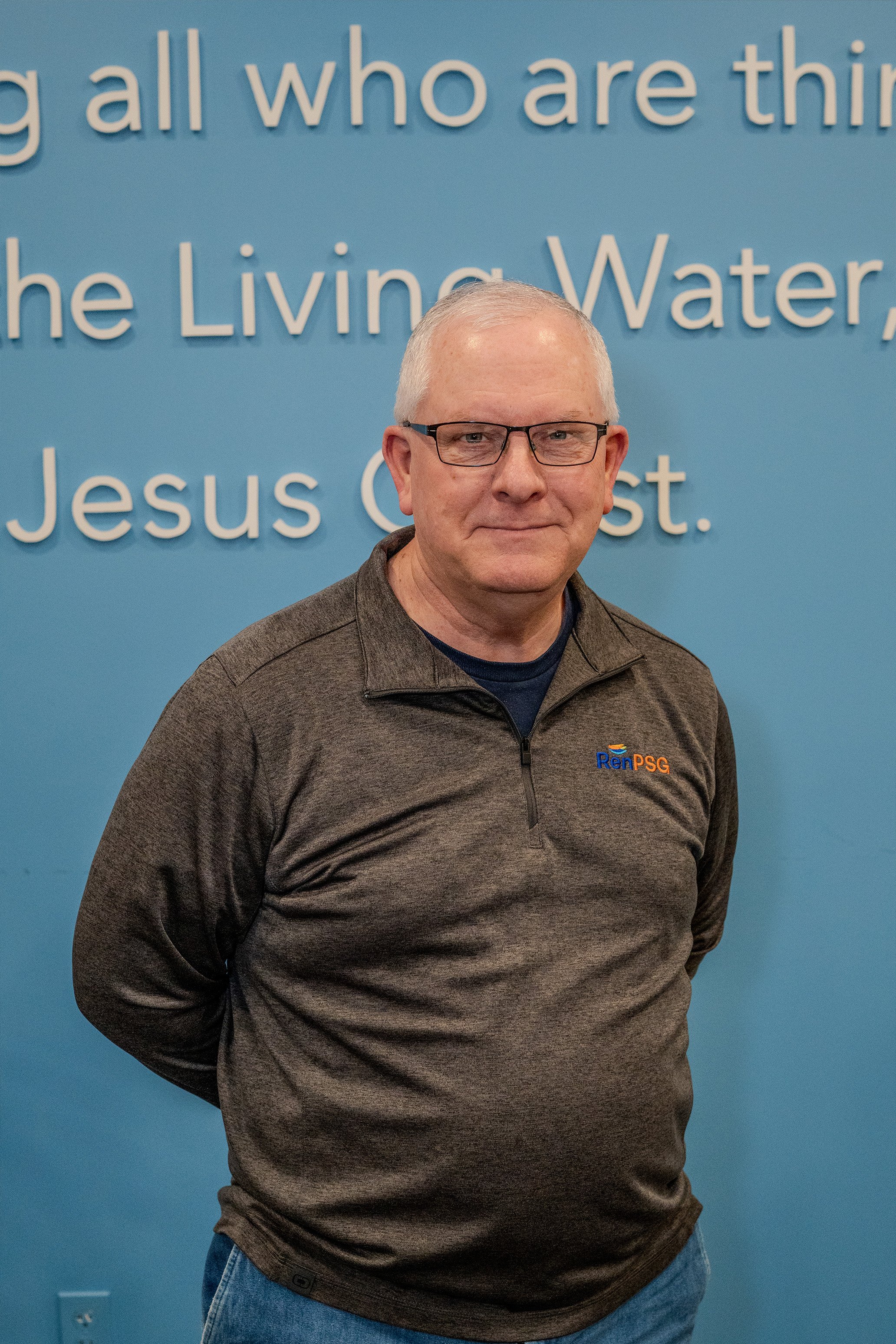 A middle-aged man with glasses, wearing a dark gray zip-up jacket with a logo, stands in front of a blue wall with white text.