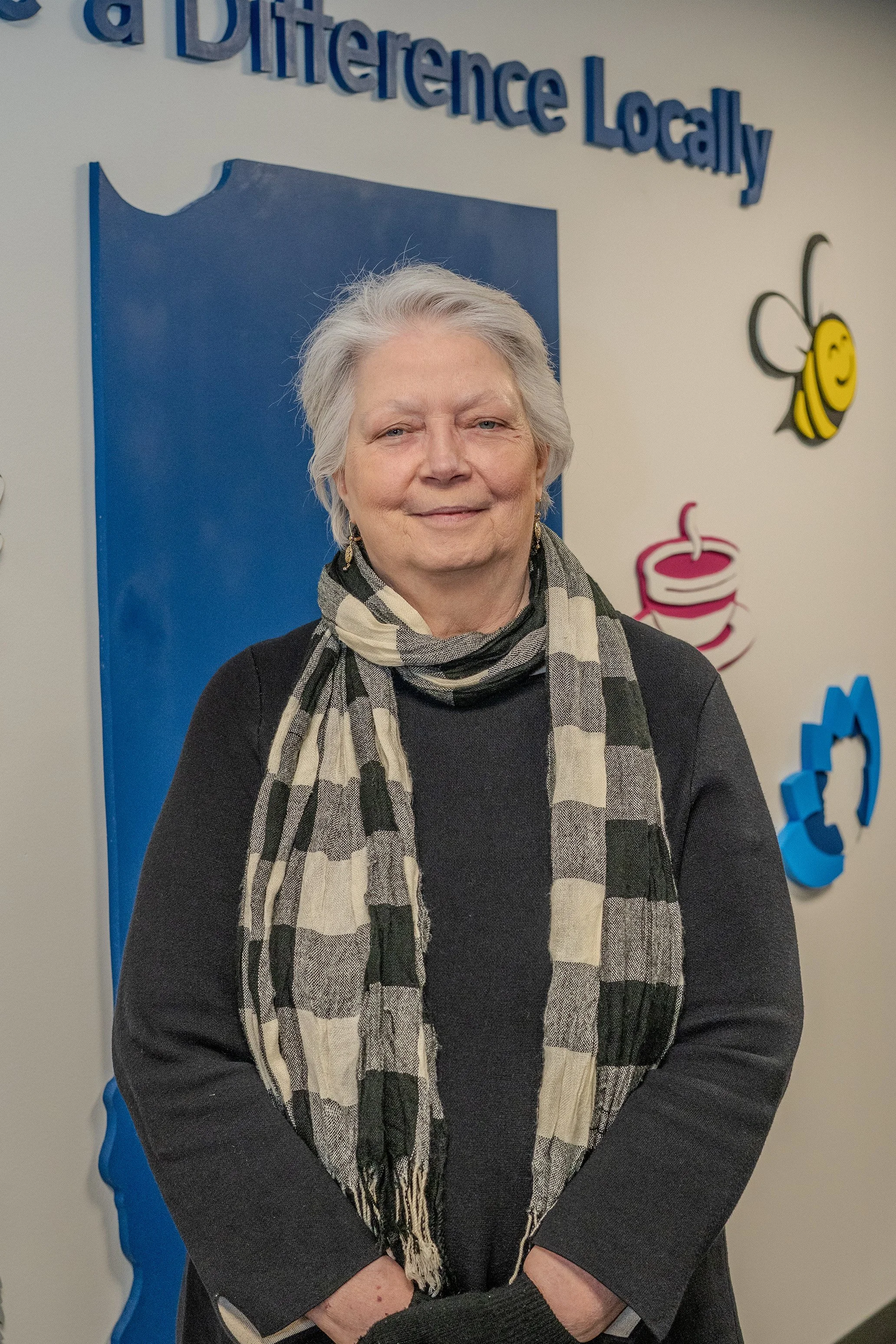 An elderly woman with gray hair, wearing a black sweater and a beige and black checkered scarf, standing in front of a wall with colorful decorations and blue 3D text that reads 'a Difference Locally'.