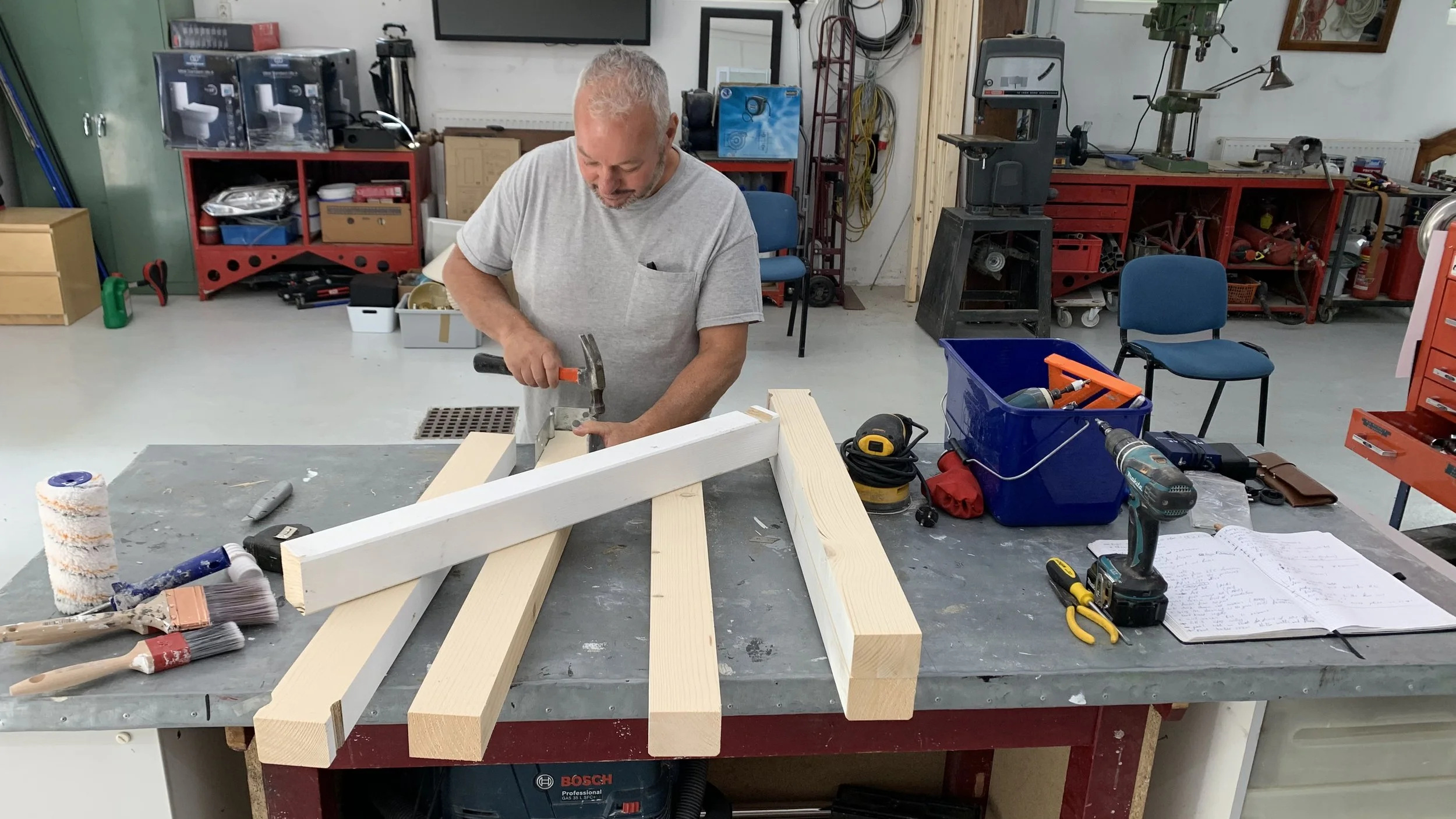 Man working in a woodshop, hammering a piece of wood on a workbench with tools and wood pieces around him.