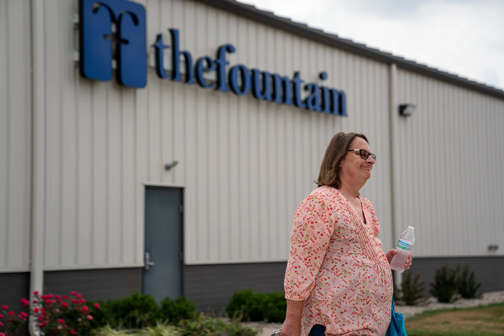 A woman with glasses walking outside a building with a sign that reads 'The FountaIn.' She is holding a water bottle and wearing a pink floral shirt.