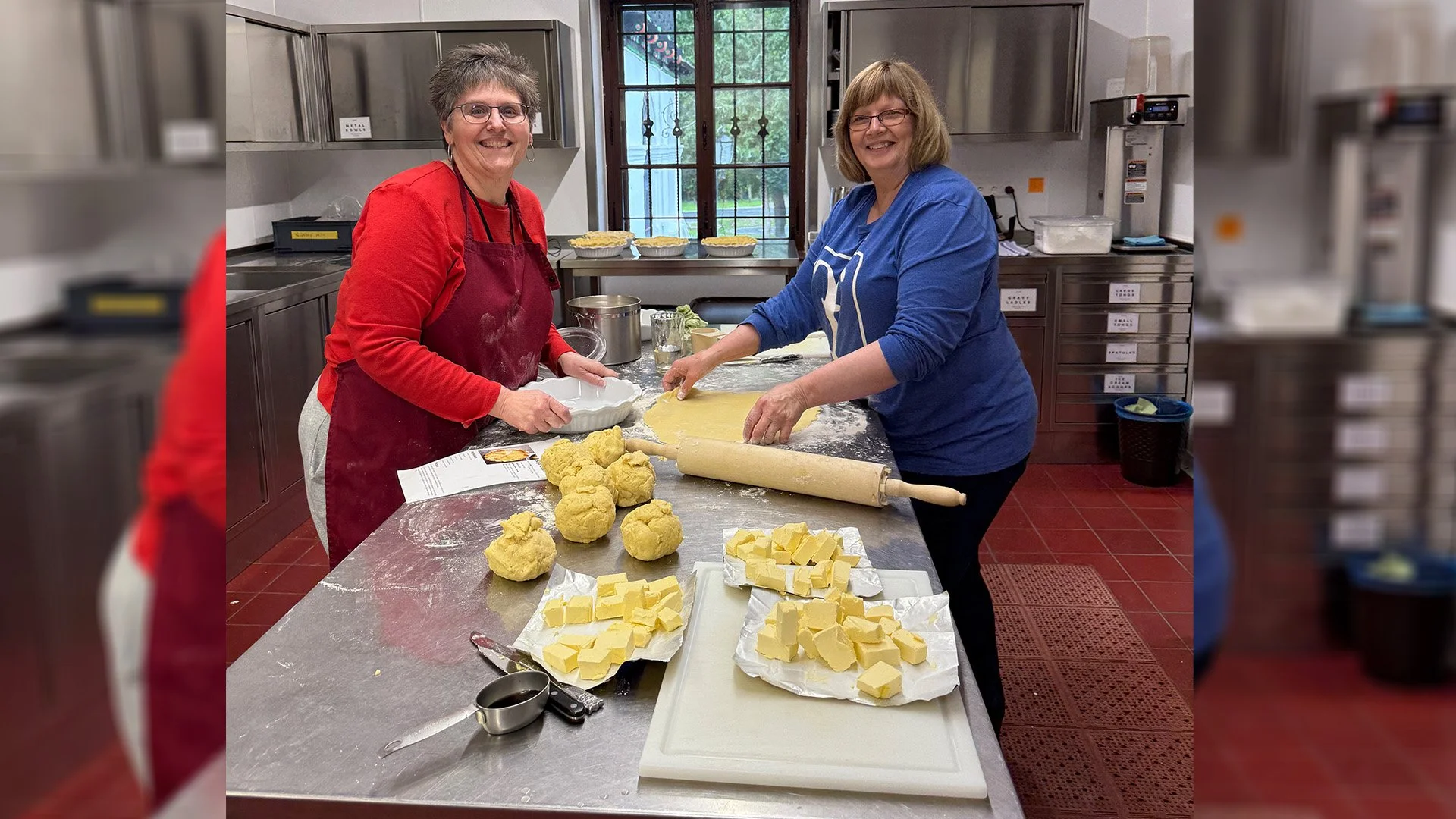 Two women baking in a kitchen, chopping and preparing butter, with dough balls on the counter and pies in the background.