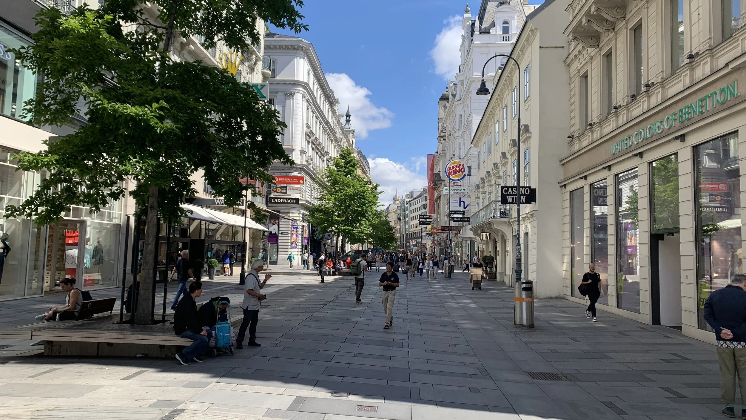 A bustling city street with people walking, sitting, and shopping, lined with white buildings and trees, with signs for Burger King and other stores, under a blue sky with some clouds.