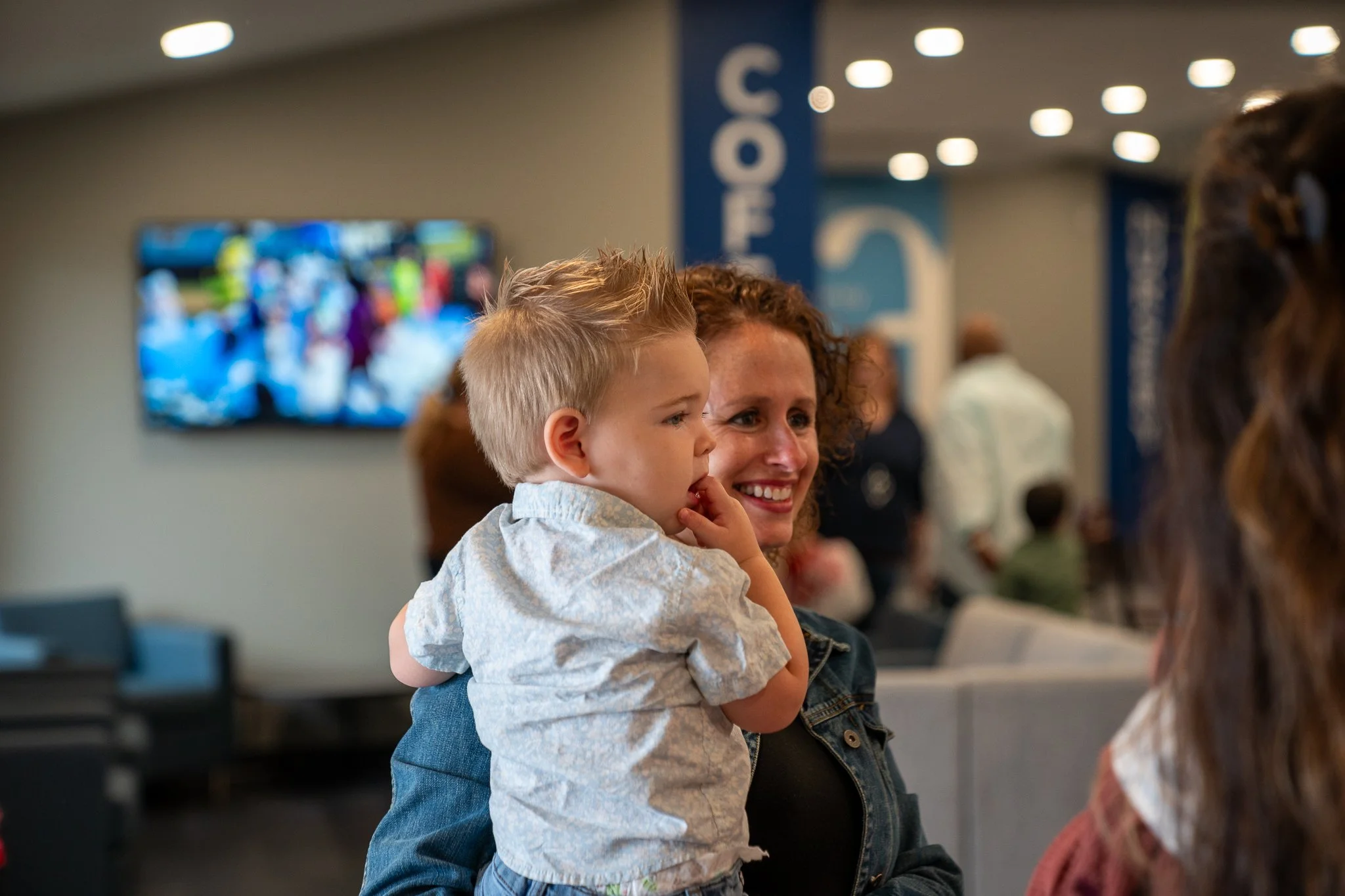 A woman holds a young boy on her lap in an airport lounge, with a television screen in the background and other travelers seated nearby.