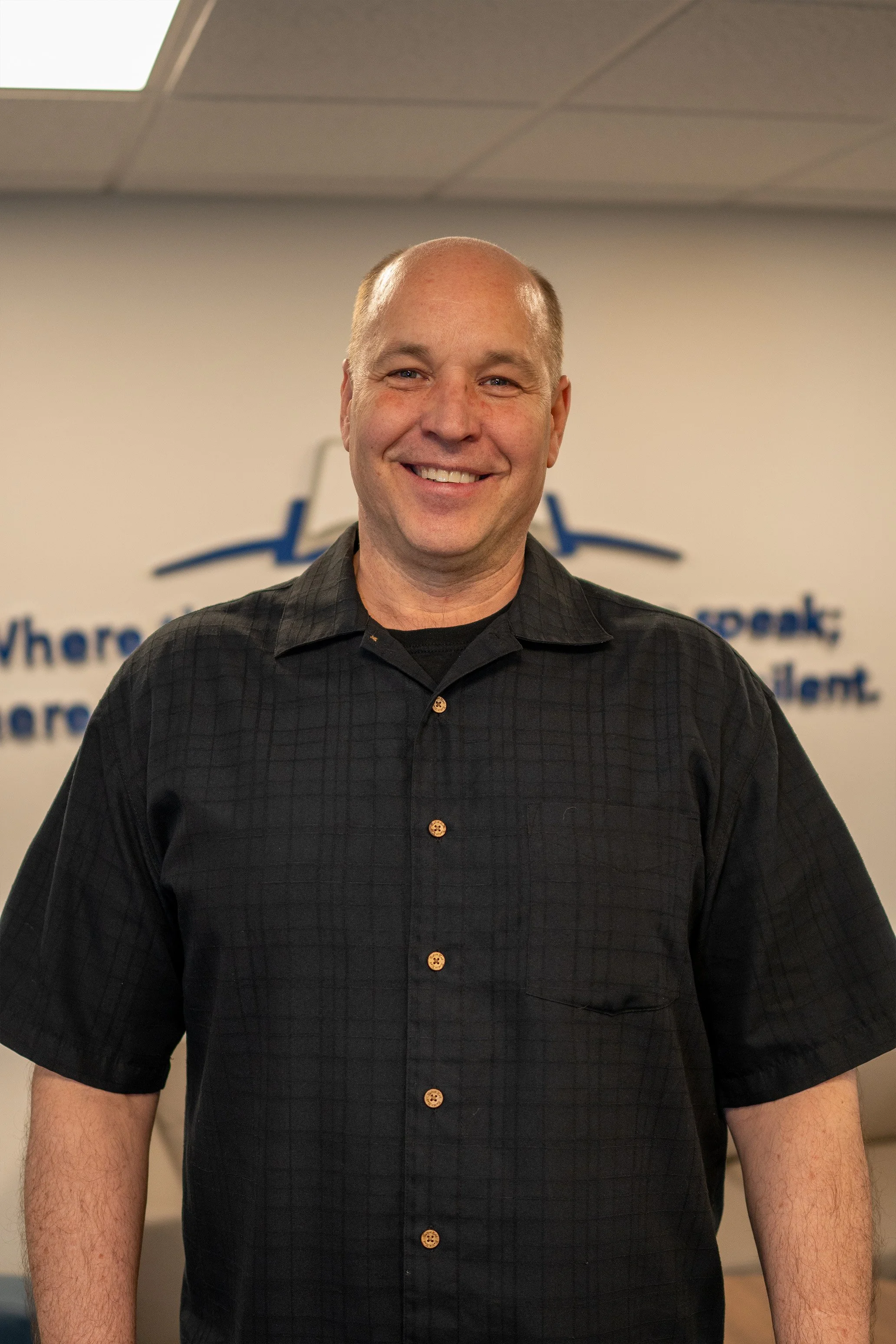 A smiling man with a shaved head wearing a black short-sleeve button-up shirt stands indoors in front of a wall with blue text and graphics.
