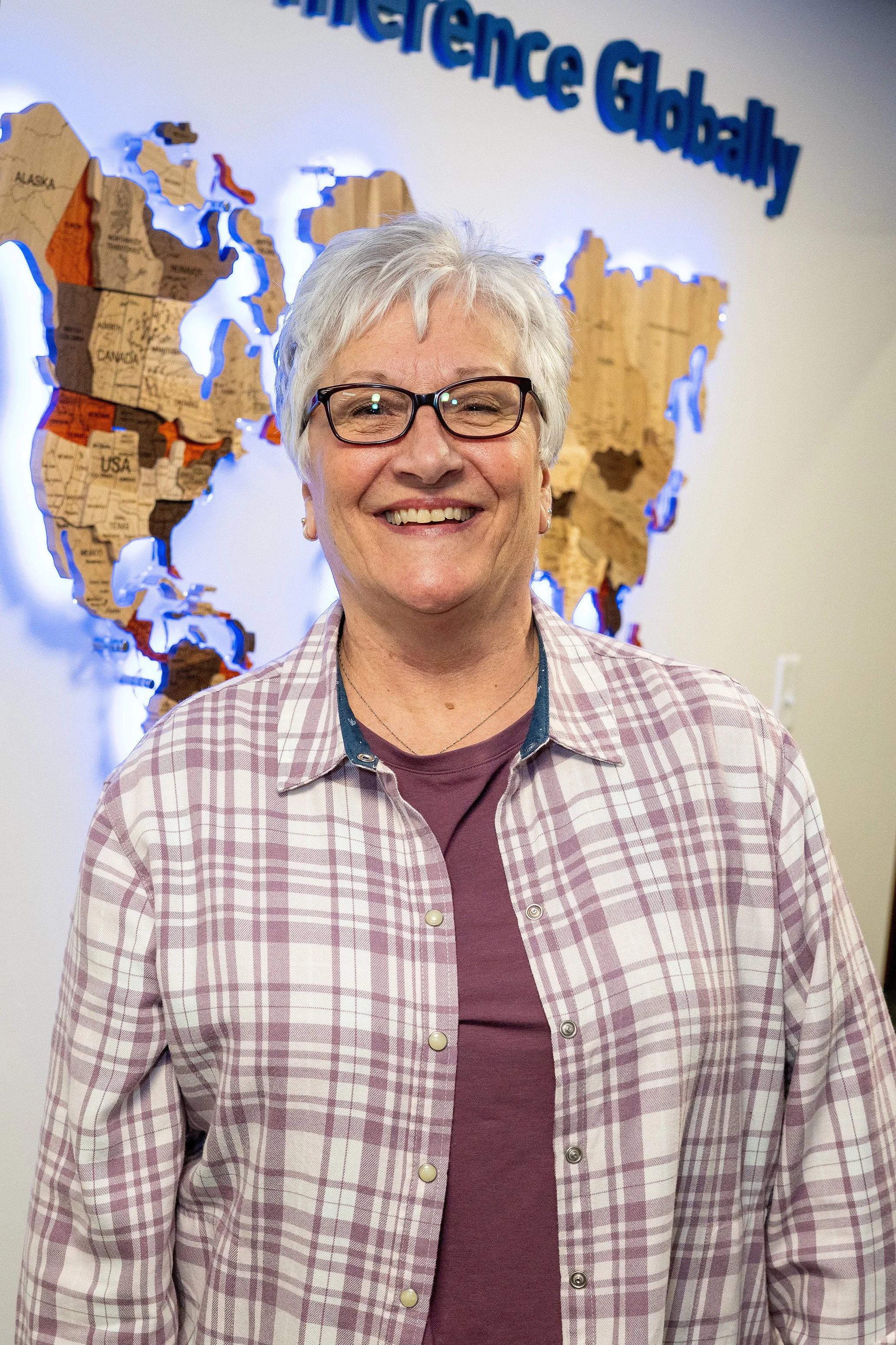 A smiling older woman with gray hair and glasses, wearing a checked shirt and a maroon t-shirt, stands in front of a map of the world with the words 'Internationally Globally' visible above.