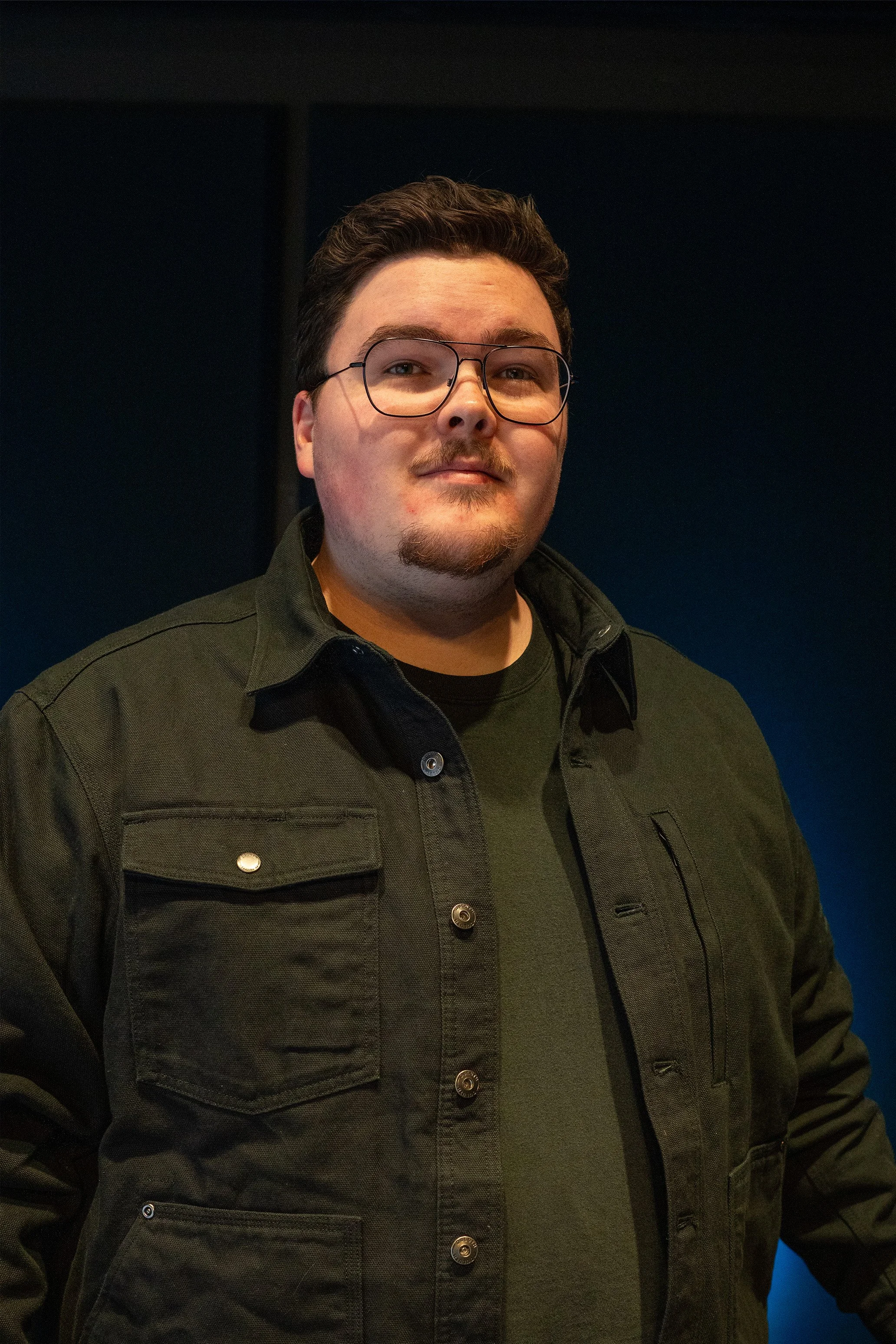 A man with glasses, a goatee, and short dark hair, wearing a dark green jacket and a black shirt, standing indoors against a dark background.