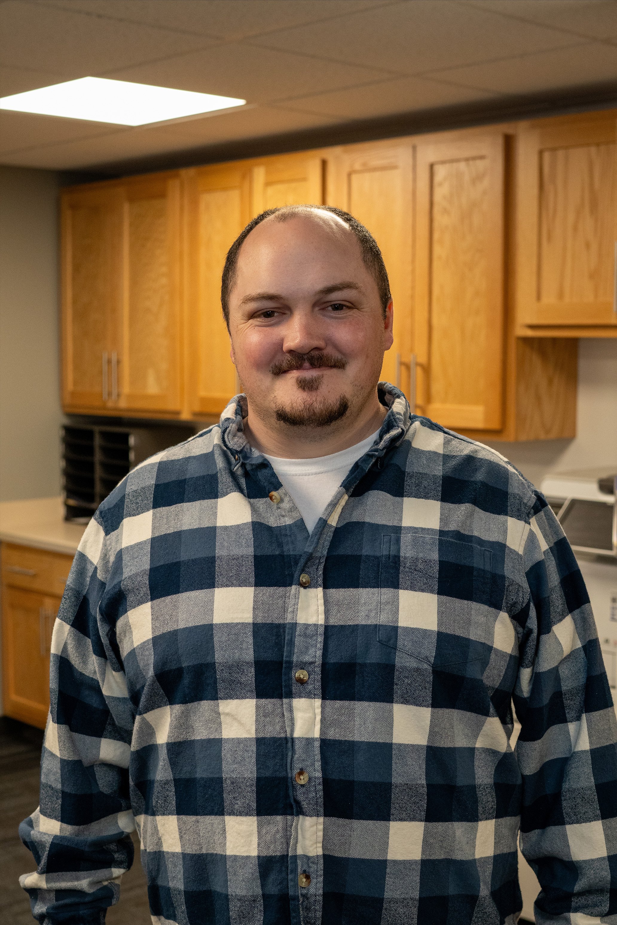 A man with short hair and a goatee, wearing a blue and white checkered flannel shirt, standing in an office or break room with wooden cabinets and fluorescent lighting.