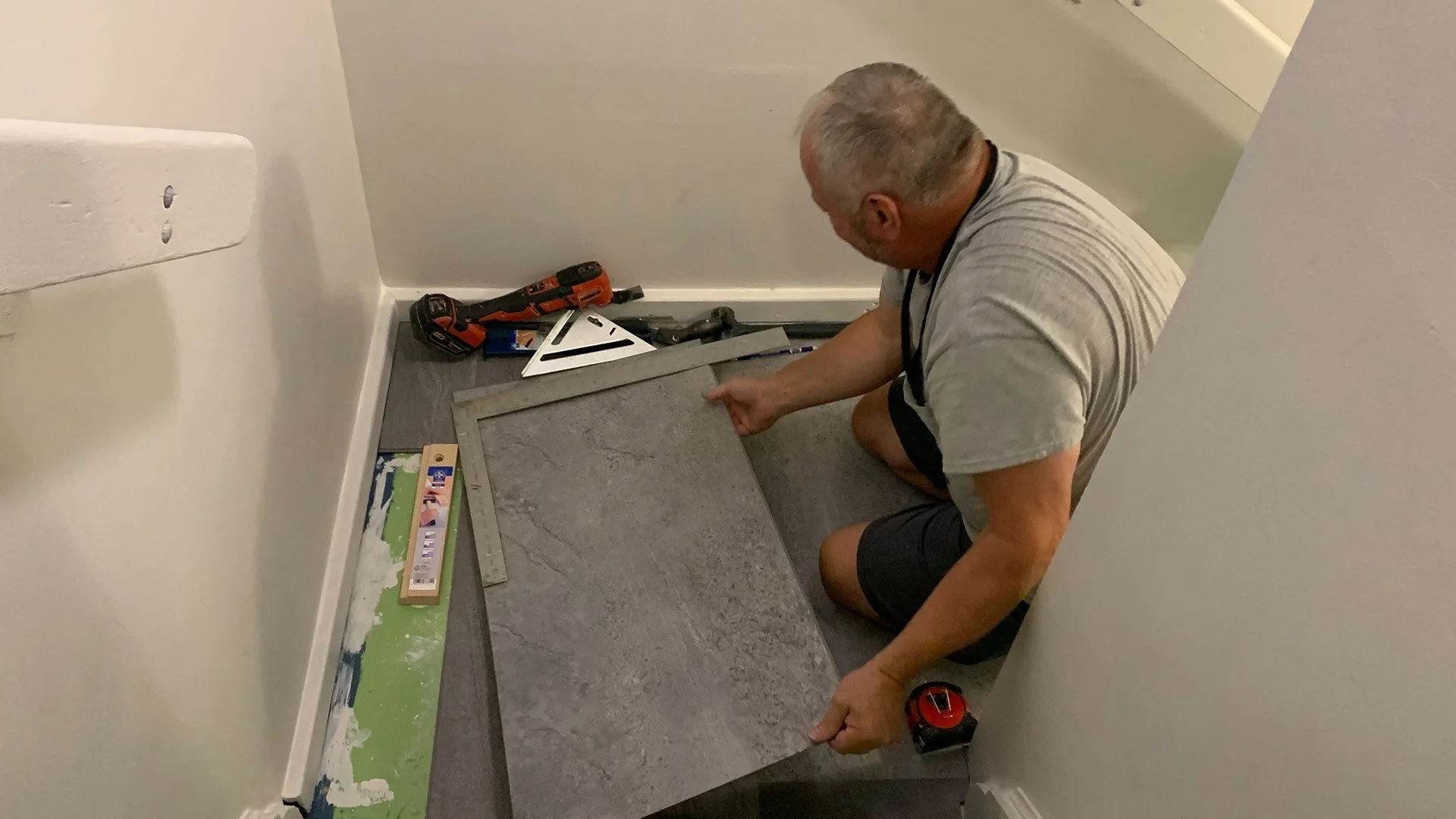 Man installing large gray ceramic tile on a floor in a corner of a room. Various tools are nearby, including a drill, a square, a measuring tape, and a level.