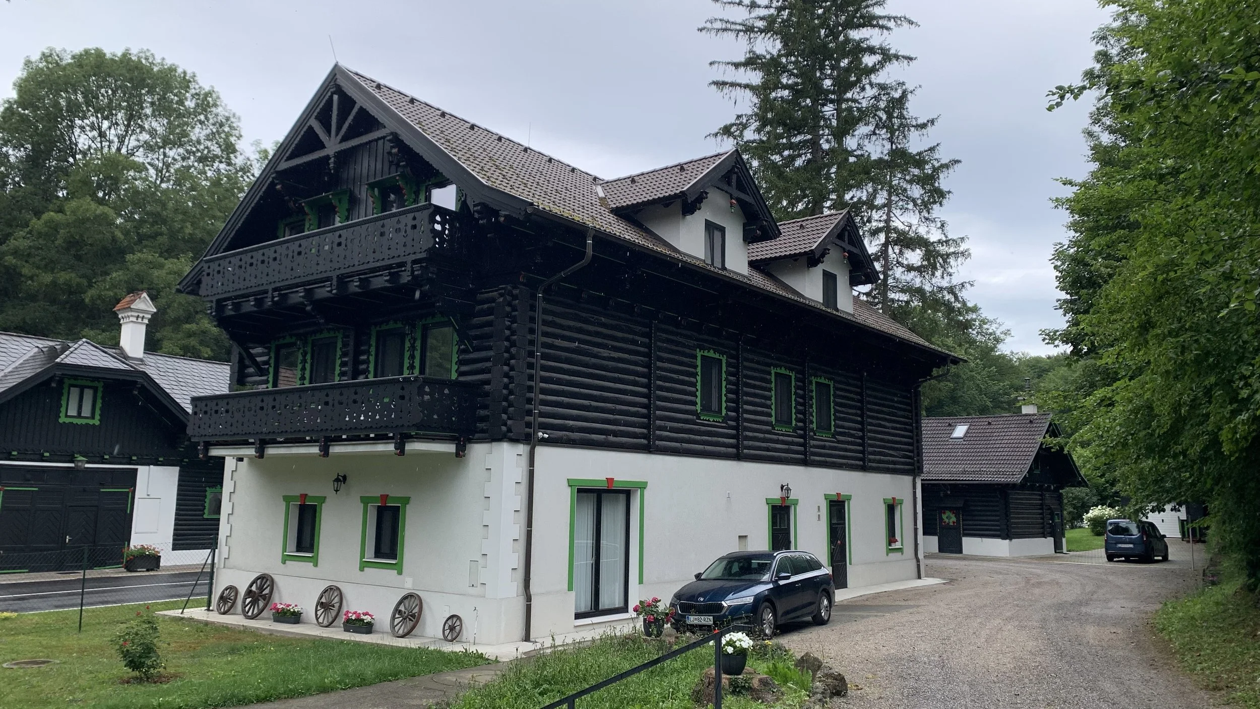 A multi-story house featuring black wood upper floors and a white lower floor with green window trims, surrounded by a garden and parked cars, with tall trees and an overcast sky in the background.