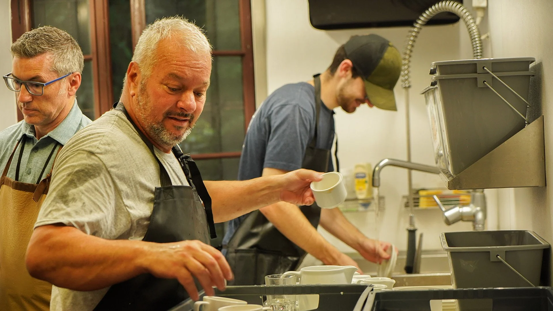 Three men washing dishes together in a kitchen, wearing aprons.