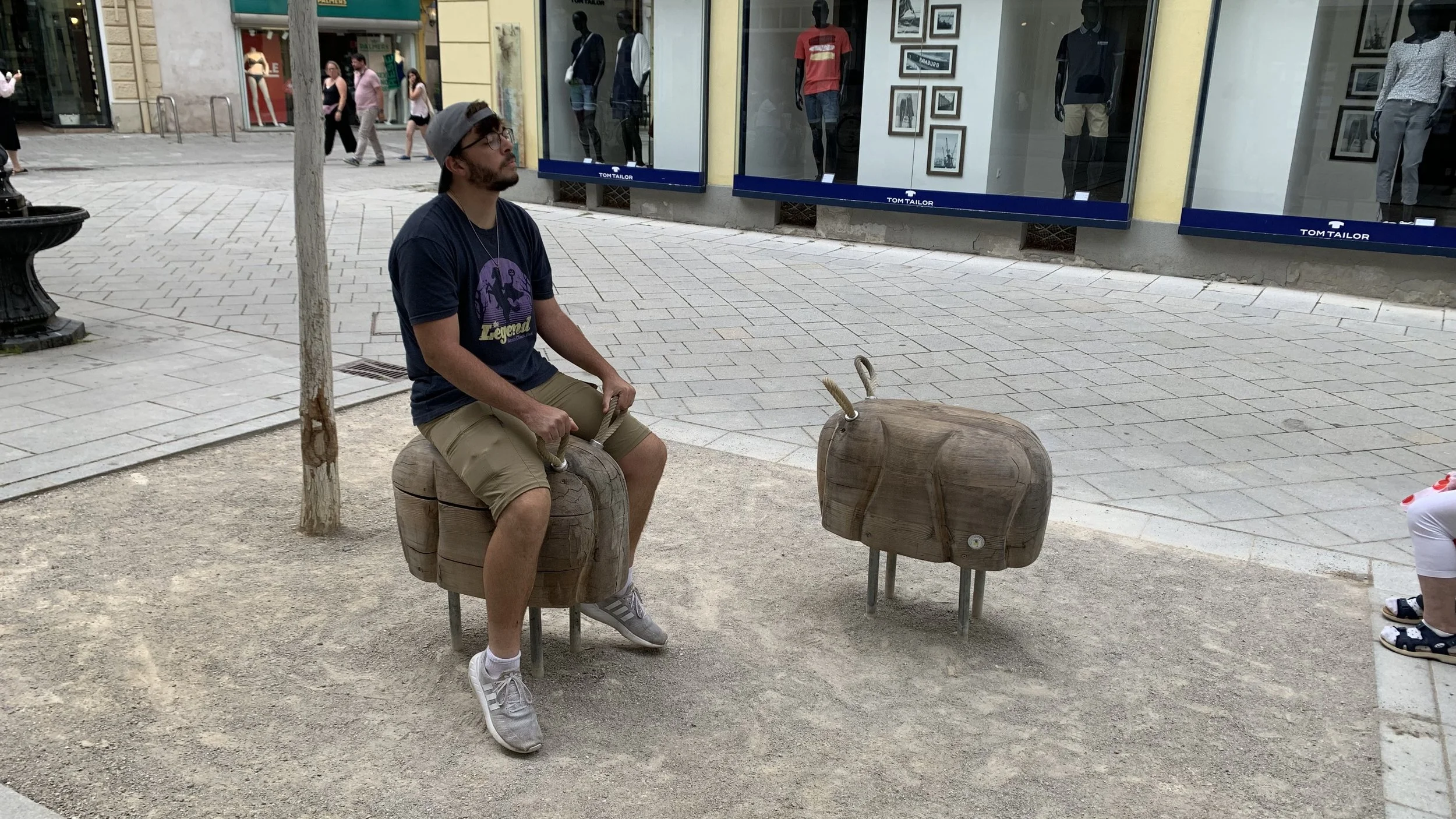 Man sitting on a wooden hippo sculpture on a city sidewalk, with storefronts and pedestrians in the background.