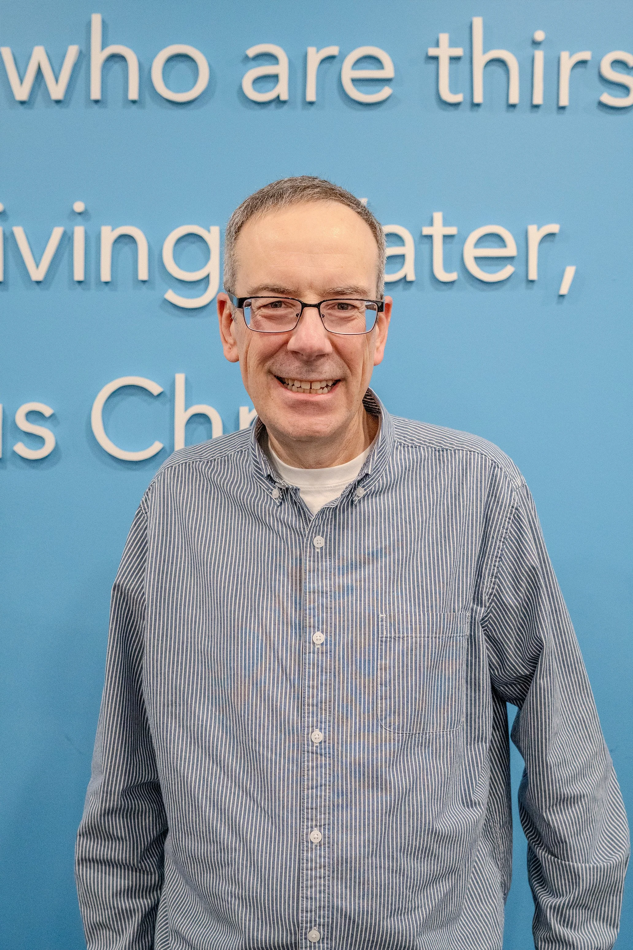 Smiling man wearing glasses and a gray striped shirt standing in front of a blue wall with partial white text.