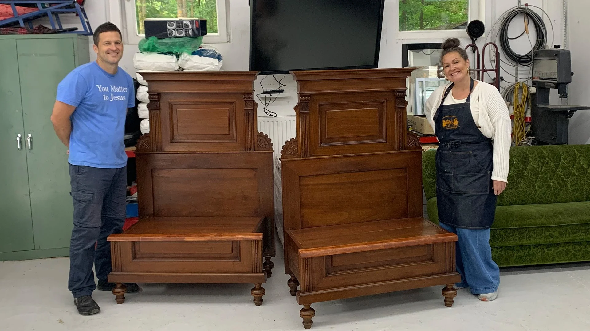 A man and a woman standing beside two matching antique wooden headboards in a workshop. The man is wearing a blue T-shirt and jeans, and the woman is wearing a white sweater, apron, and jeans. They are smiling at the camera. The workshop has various