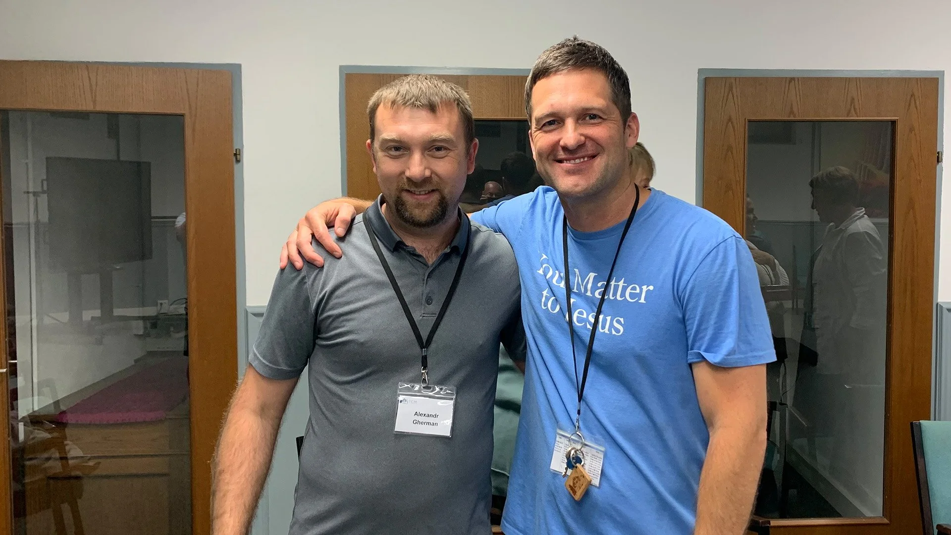 Two smiling men standing close together with arms around each other's shoulders in an indoor room with wooden framed glass doors in the background. One man is wearing a gray polo shirt and the other a blue T-shirt that reads 'You Matter to Jesus'.