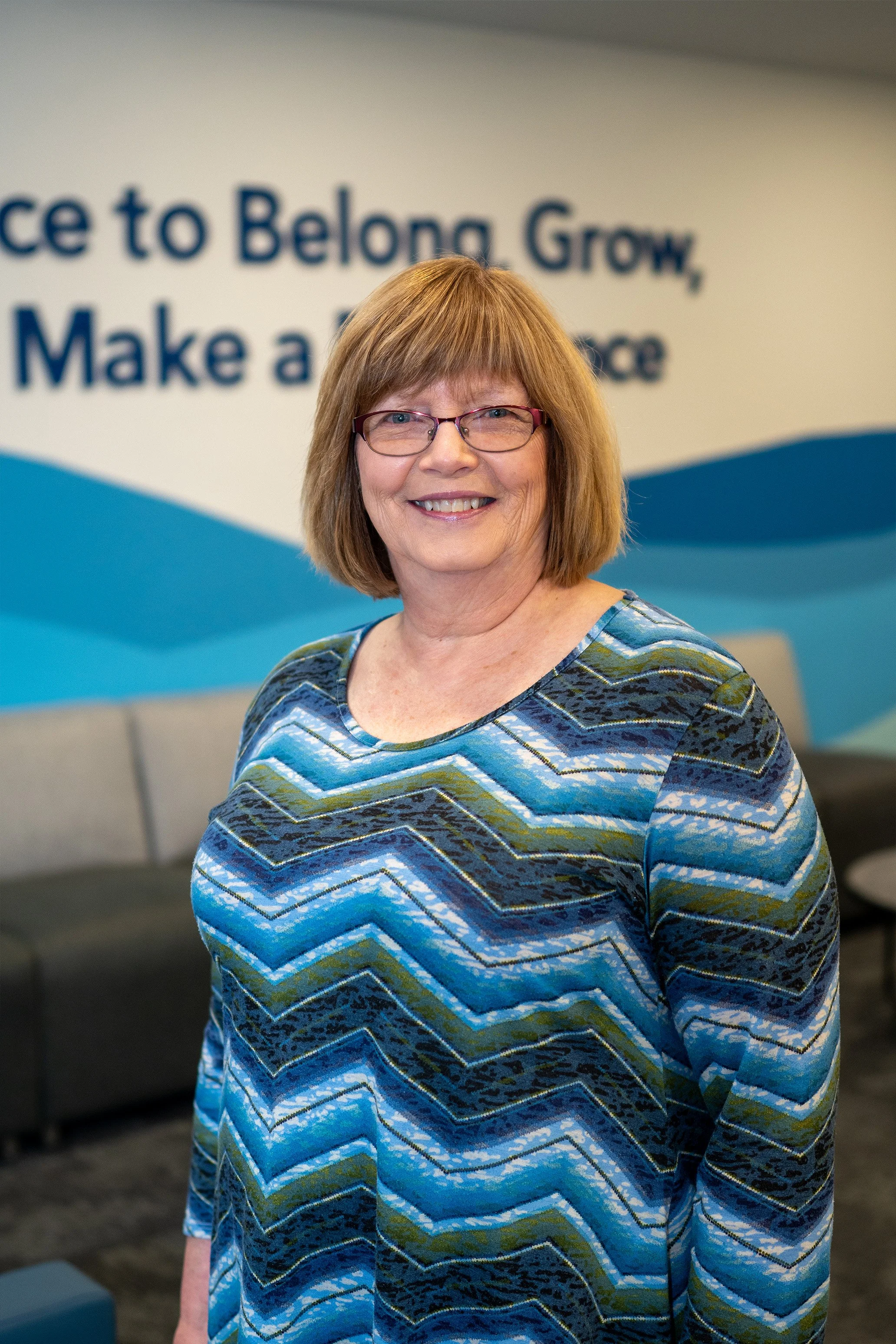 A smiling woman with glasses and shoulder-length reddish hair standing indoors in front of a sign with partially visible text. She is wearing a blue patterned long-sleeve shirt.