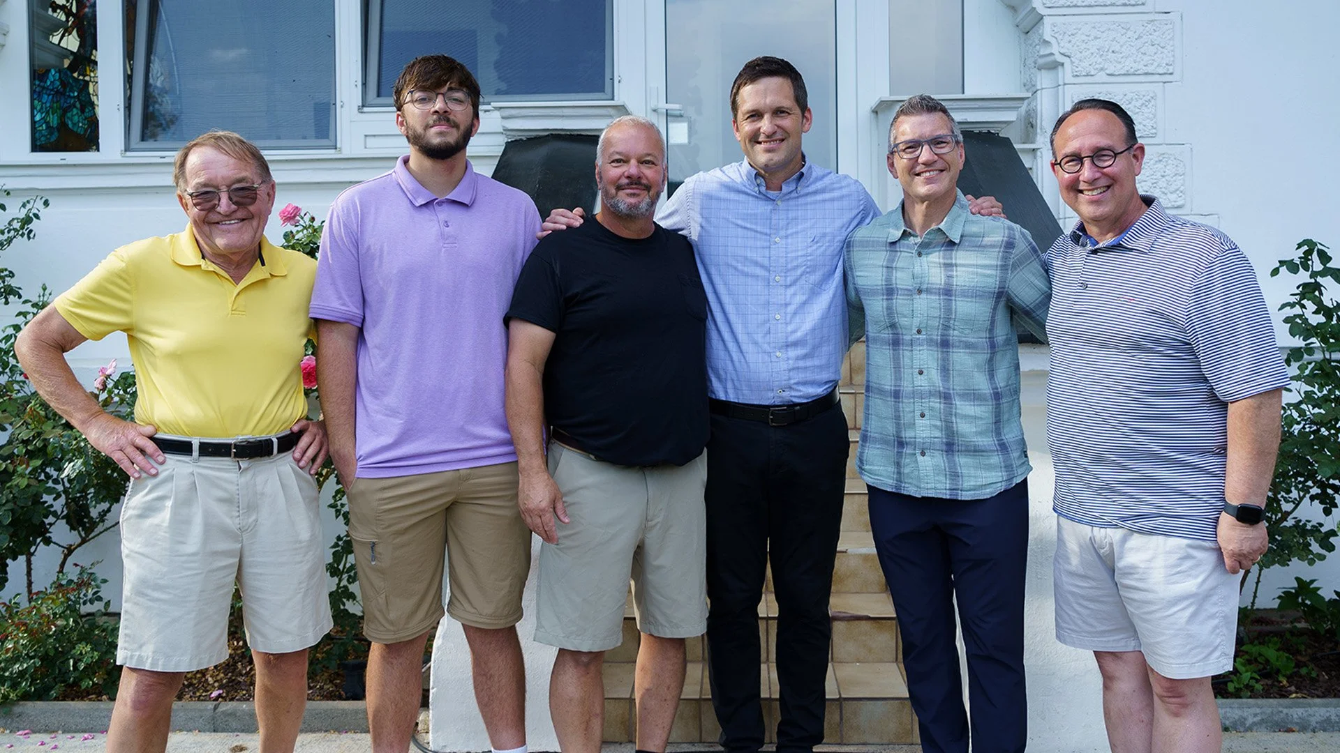 Group of seven men standing outside in front of a house, smiling and posing for a photo.