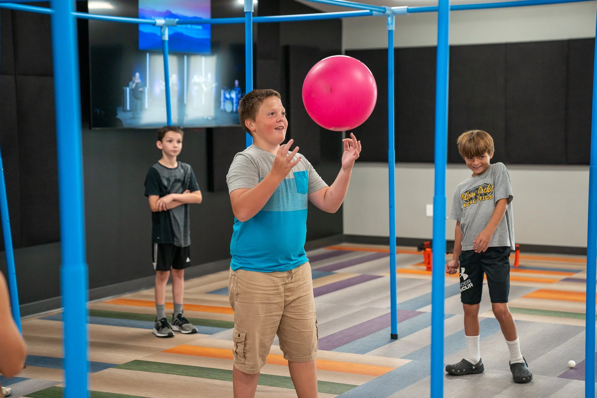Children playing with a pink ball in an indoor activity room with colorful carpet and play structure.