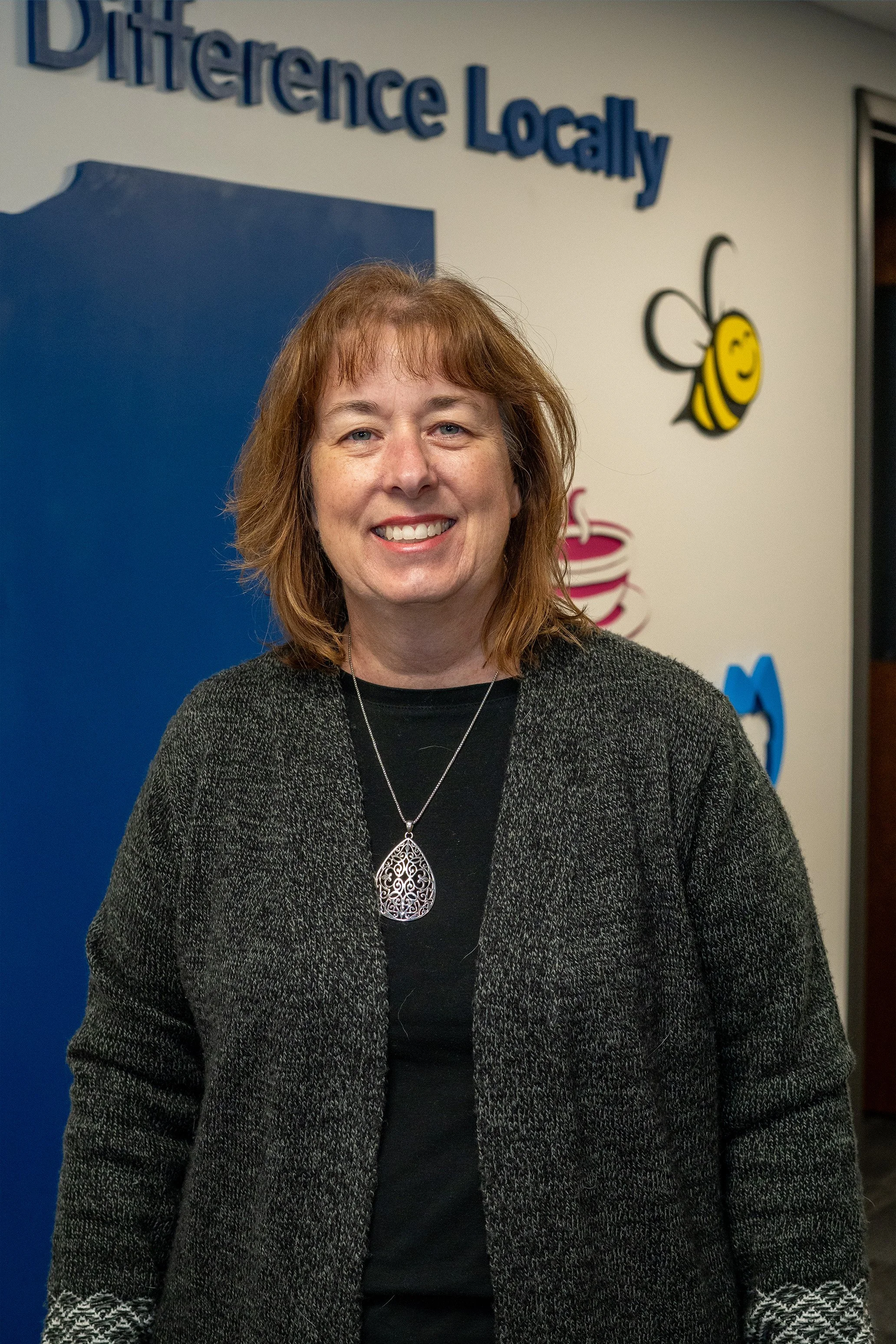 A woman with shoulder-length light brown hair smiling, standing in front of a wall with colorful decorations, including a bee, a speech bubble, and the partial text "Difference Locally."