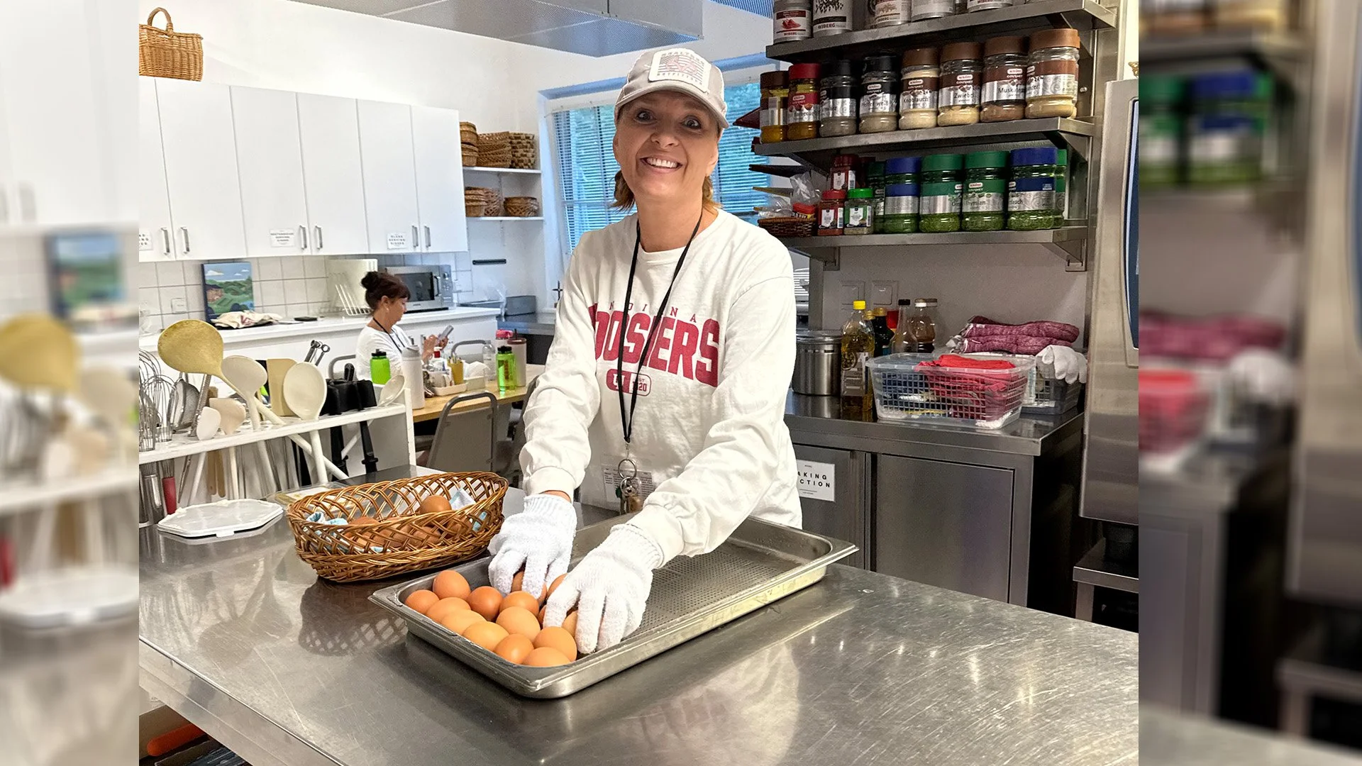 A woman smiling and wearing a gray cap and white sweatshirt with red lettering, standing at a stainless steel table, placing eggs into a tray in a commercial kitchen. Another person is working in the background, surrounded by kitchen supplies and she