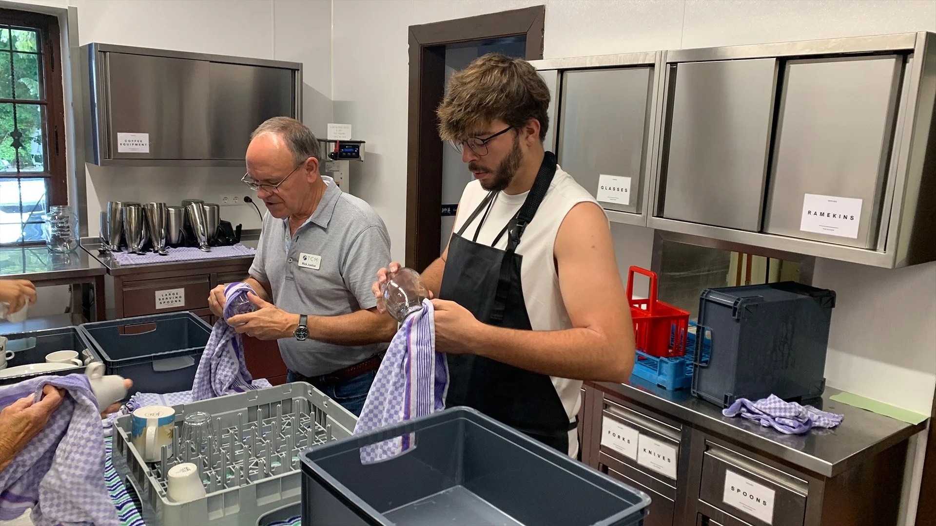 Two men, one older and one younger, are washing dishes at a sink in a kitchen or dishwashing station. They are using purple checkered dish towels. There are labeled cabinets, and various kitchen items and dishware around them.