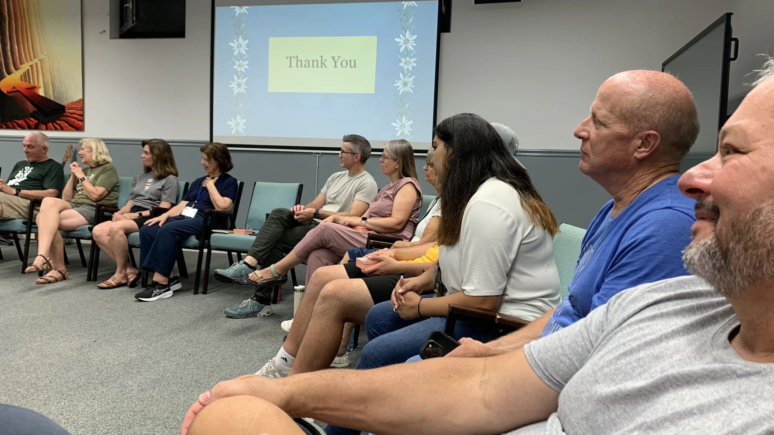 Group of people seated in a conference room, facing a screen displaying a tribute slide with the words 'Thank You' and decorative flowers.
