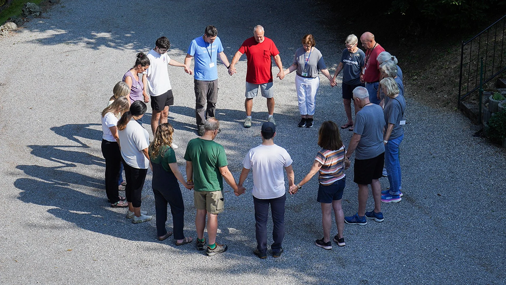 A group of around 20 people holding hands in a circle outdoors on a gravel surface, participating in a group activity or prayer on a sunny day.