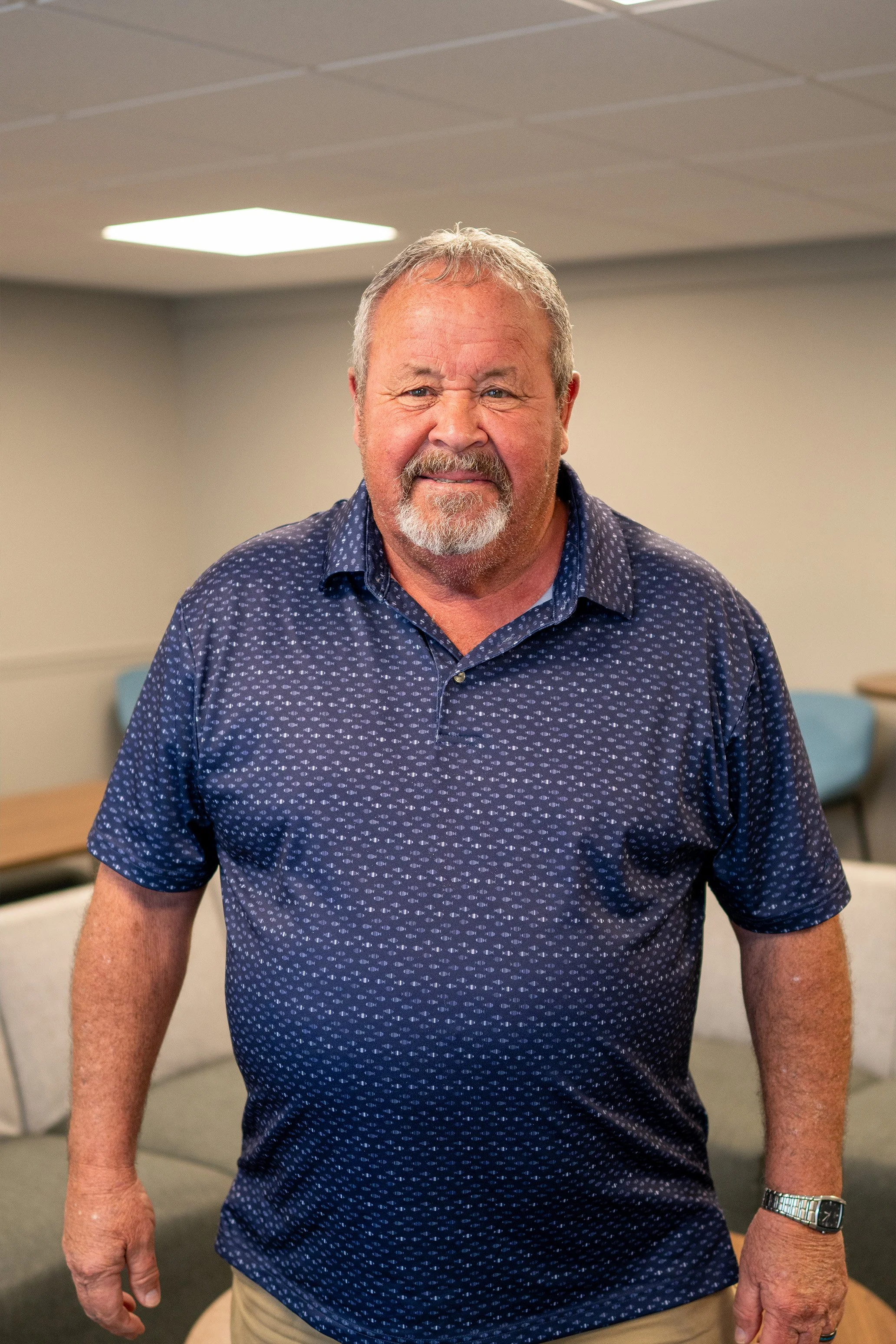 A smiling middle-aged man with a beard and short hair, wearing a navy blue patterned polo shirt and tan pants, standing in a room with beige walls and a ceiling with fluorescent lighting.