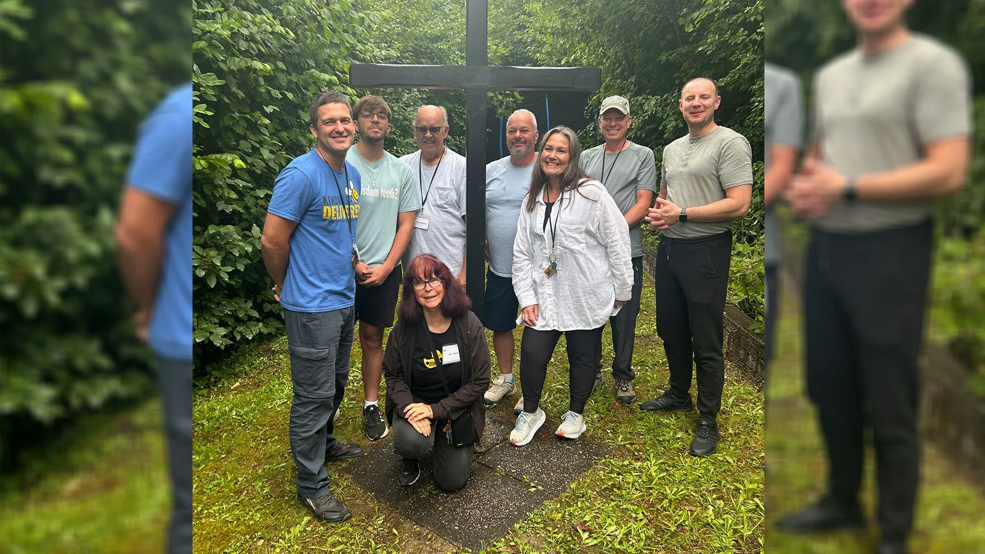 Group of nine people standing outdoors on a trail, posing in front of a large black wooden cross surrounded by green foliage.