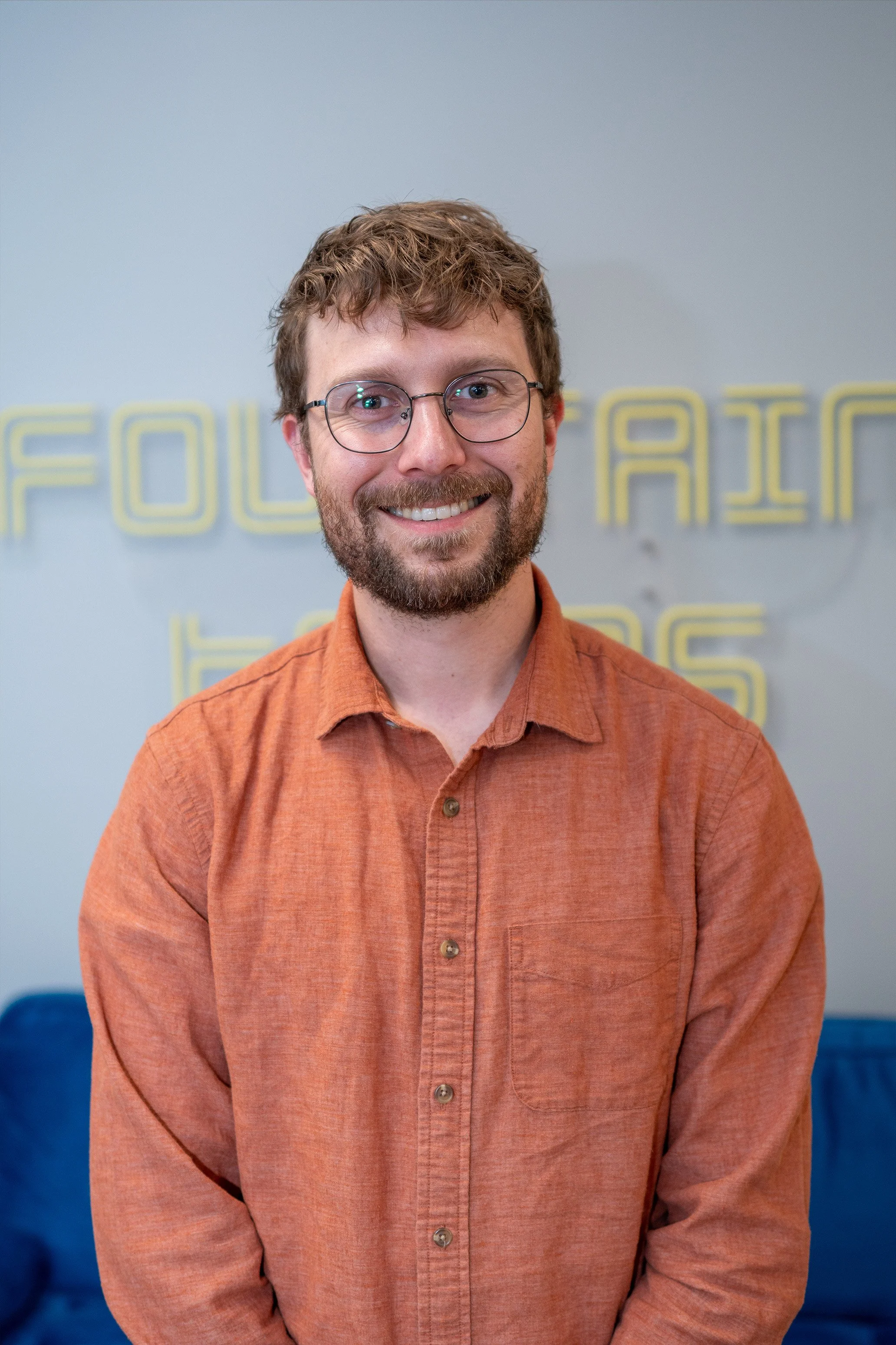 A smiling man with glasses and a beard wearing an orange shirt, standing indoors in front of a wall with yellow illuminated text. The background includes blue seating and a wall with glowing yellow letters.