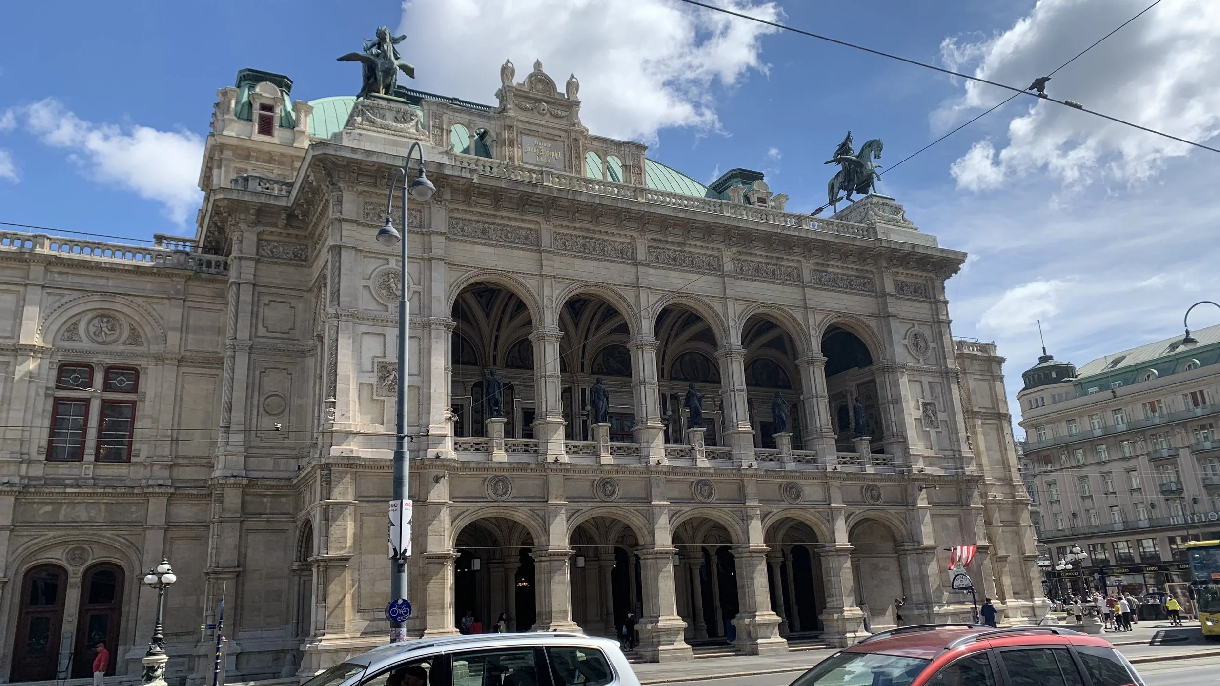 A historic building with ornate architecture, statues in niches, and green domed rooftop with two large bronze statues of horses and riders on top, under a partly cloudy sky.