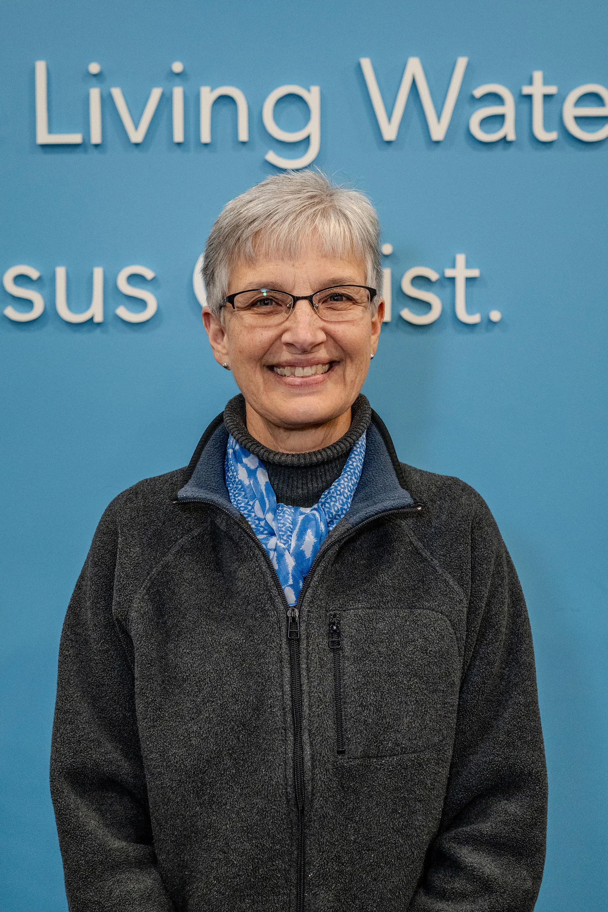 A smiling woman with short gray hair, glasses, and wearing a dark gray fleece jacket over a black turtleneck and blue patterned scarf, standing in front of a blue wall with white text that reads 'Living Water'.