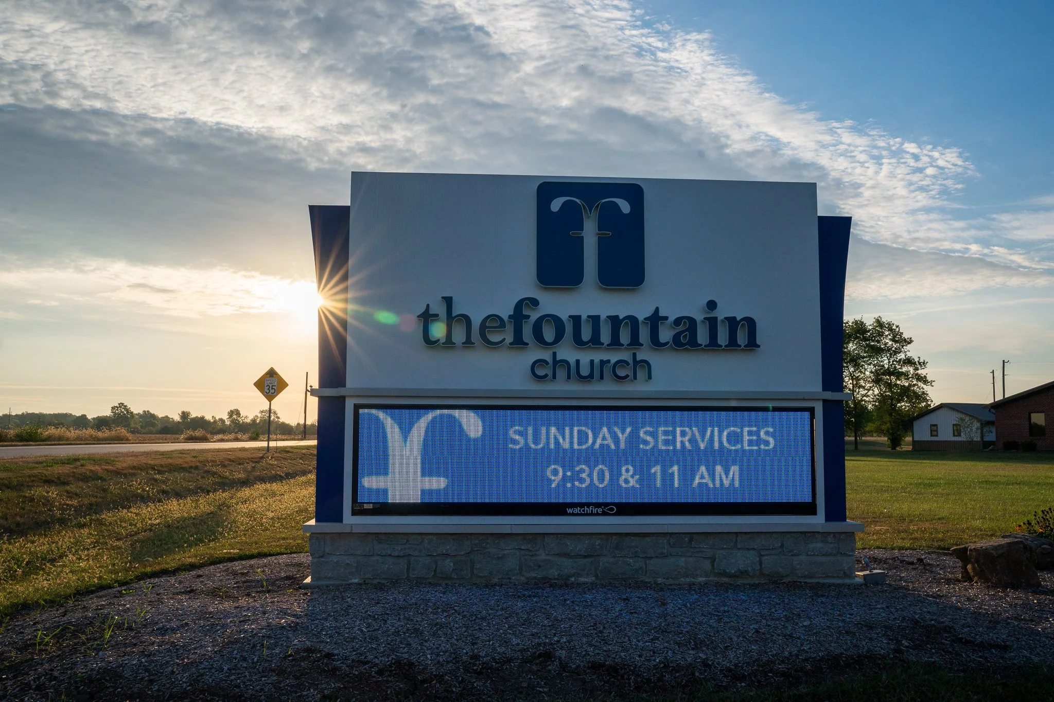 Sign for The Fountain Church with service times displayed, set against a background of a partly cloudy sky and sunset.
