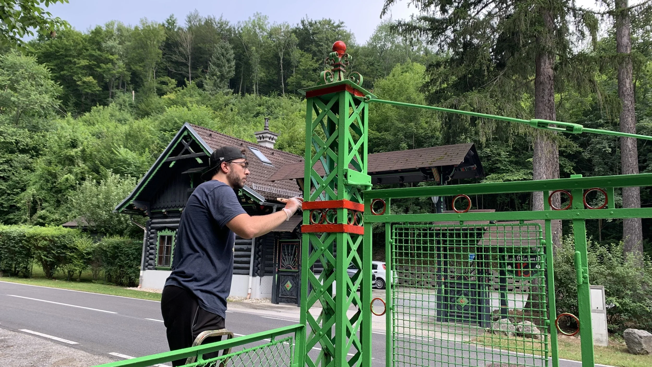 Young man wearing glasses and a black cap installing a green metal structure on a roadside with trees and a house in the background.