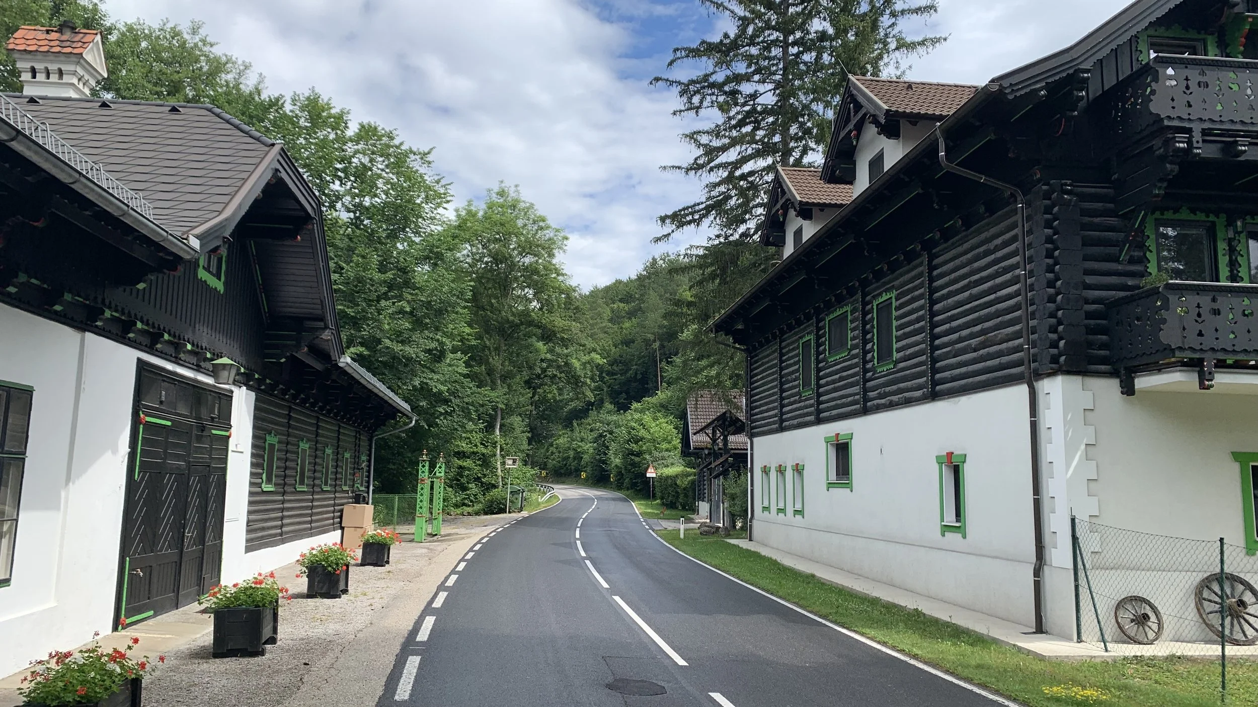 A quiet, winding country road flanked by houses with black and white exteriors, green window frames, and brown roofs, surrounded by lush green trees under a partly cloudy sky.