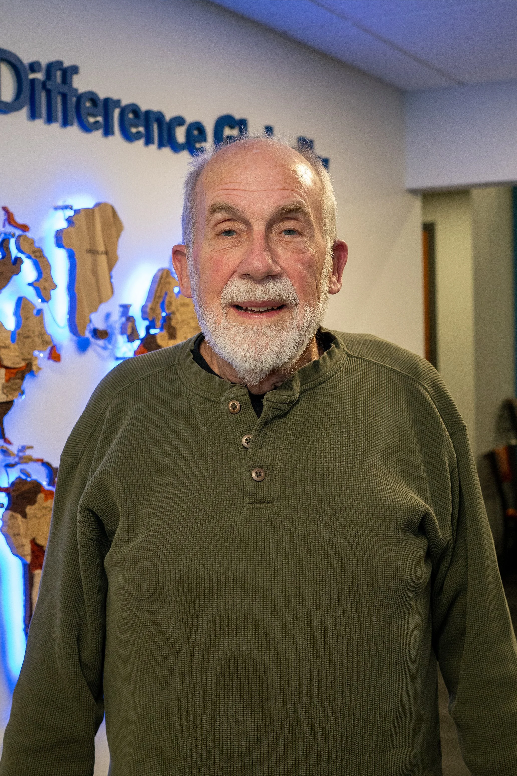An older man with a white beard and grey hair smiling, standing in front of a wall with a map of the world and the words 'Difference of...' partially visible.