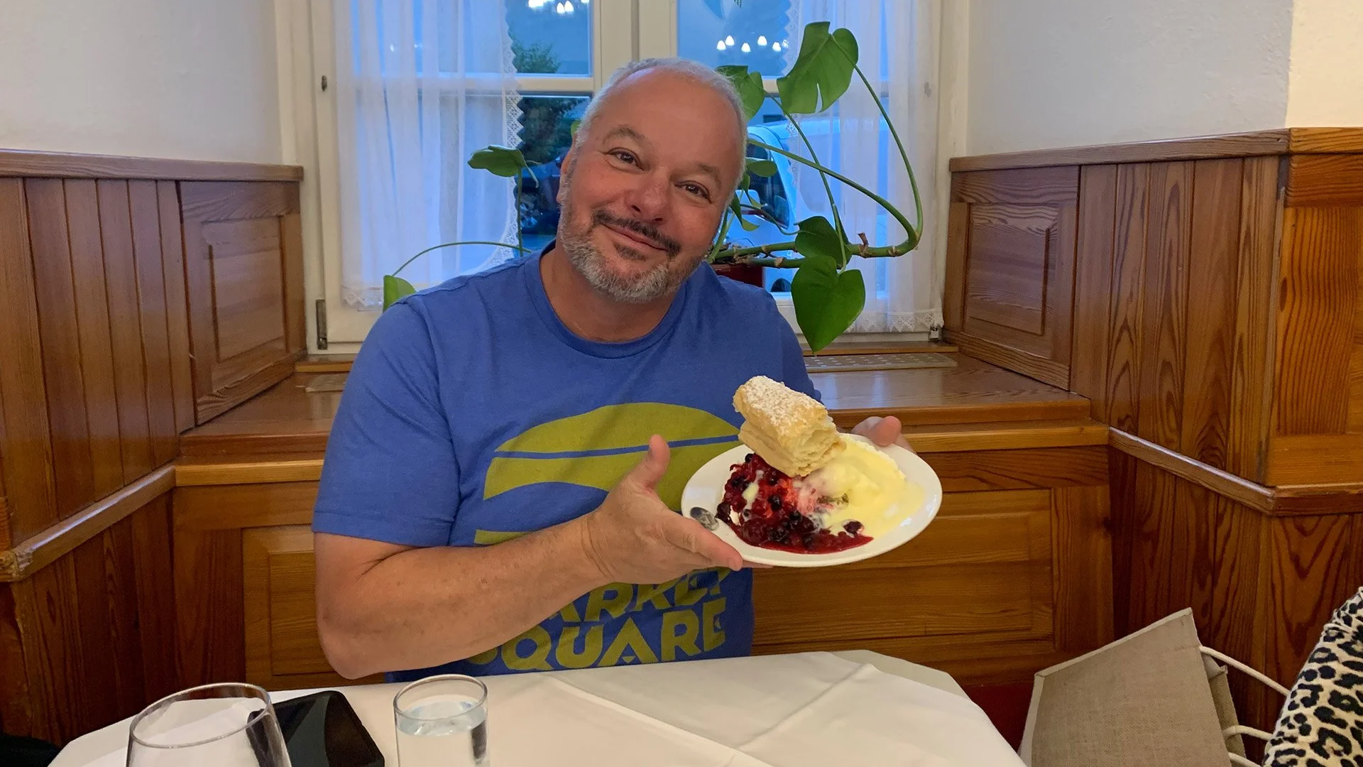 A smiling man with a beard in a blue t-shirt holding a plate with dessert, including a pastry, berries, and whipped cream, seated at a table in a cozy restaurant with wooden paneling and a window with lace curtains.