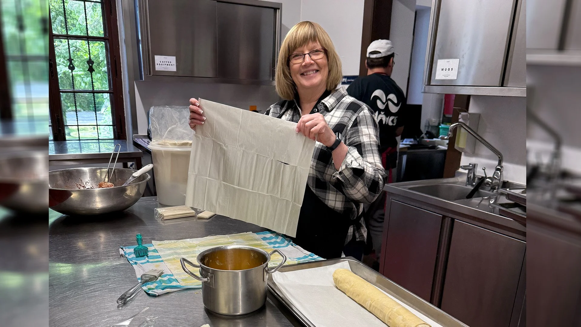 A smiling woman holding a sheet of puff pastry in a kitchen with baking tools, a rolling pin, and a baking sheet with a rolled pastry.