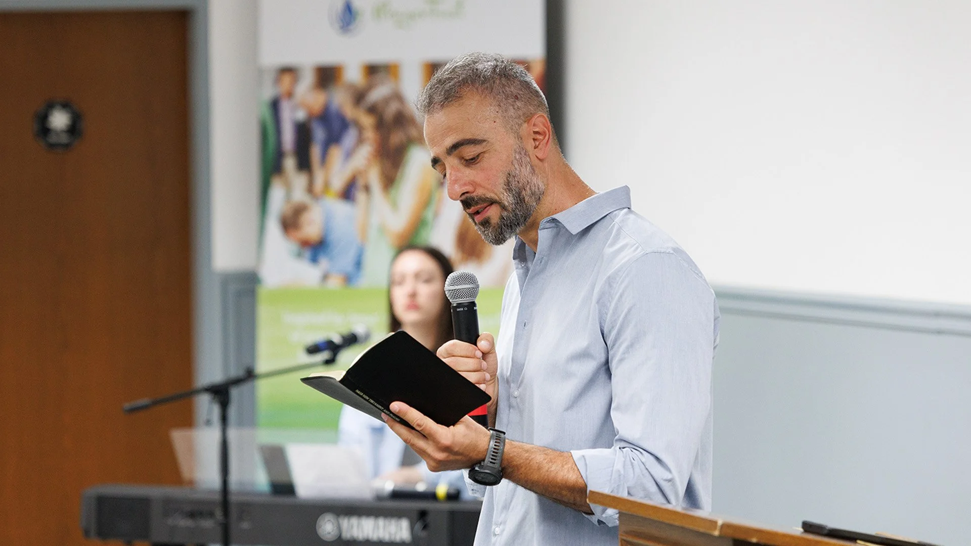 A man with gray hair and beard holding a microphone and reading from a black booklet in a conference room. A woman is sitting in the background at a keyboard with a blurred poster on the wall behind them.