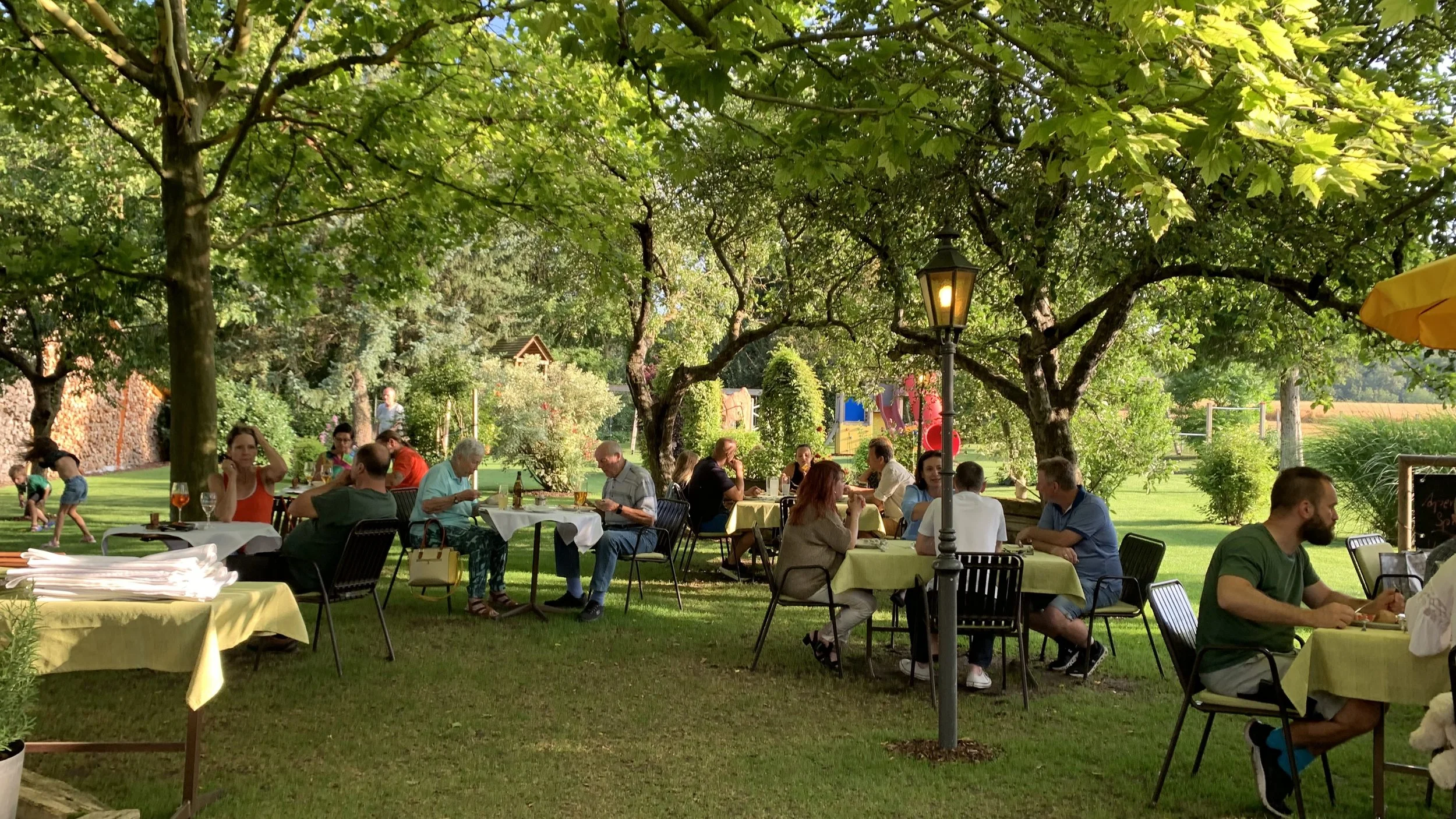Outdoor restaurant with people dining at tables under large trees, with a lamp post in the center and children playing in the background.