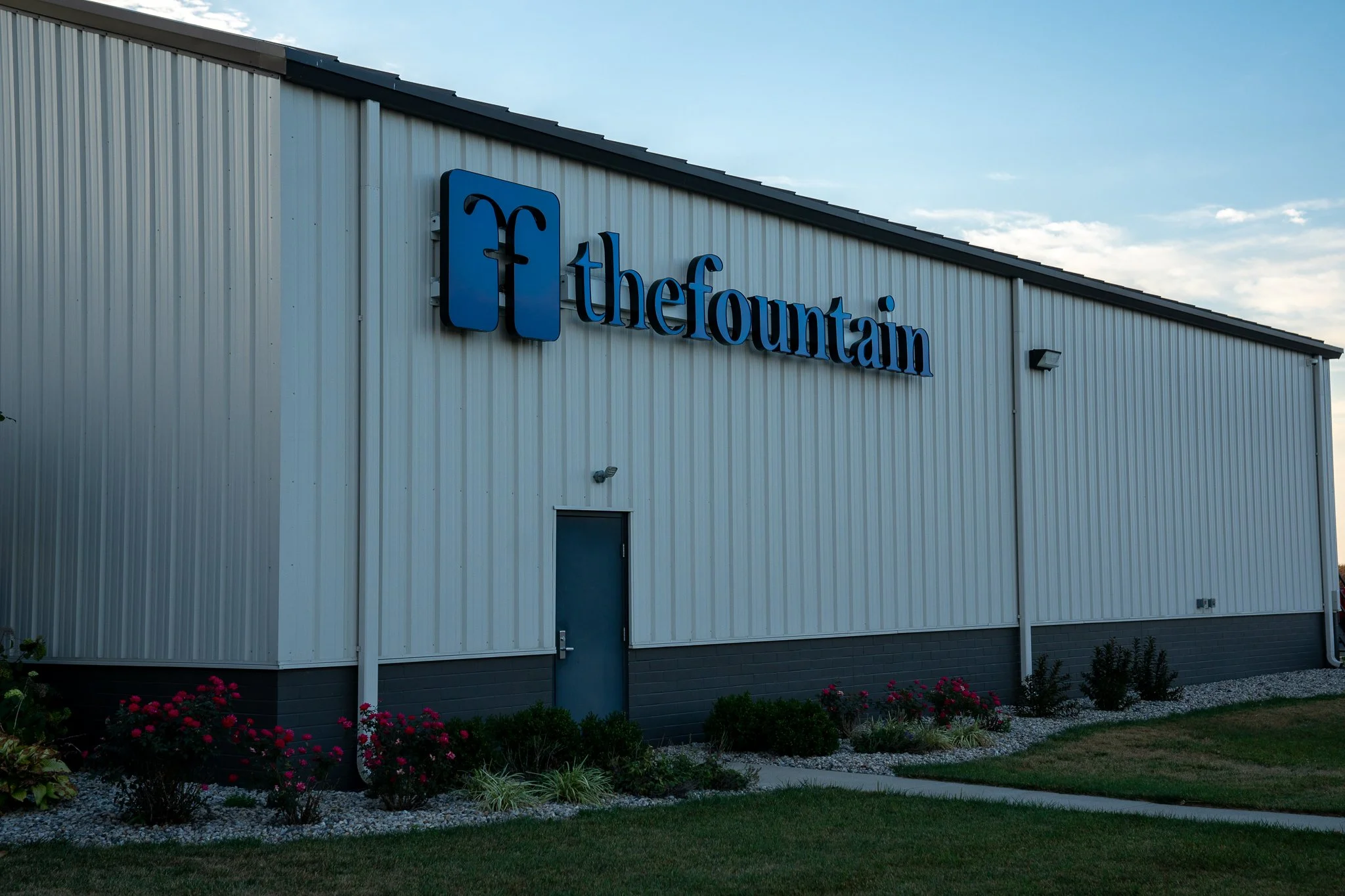 Building with a sign that reads 'the fountain' and has the Facebook logo, surrounded by green grass and flowers.