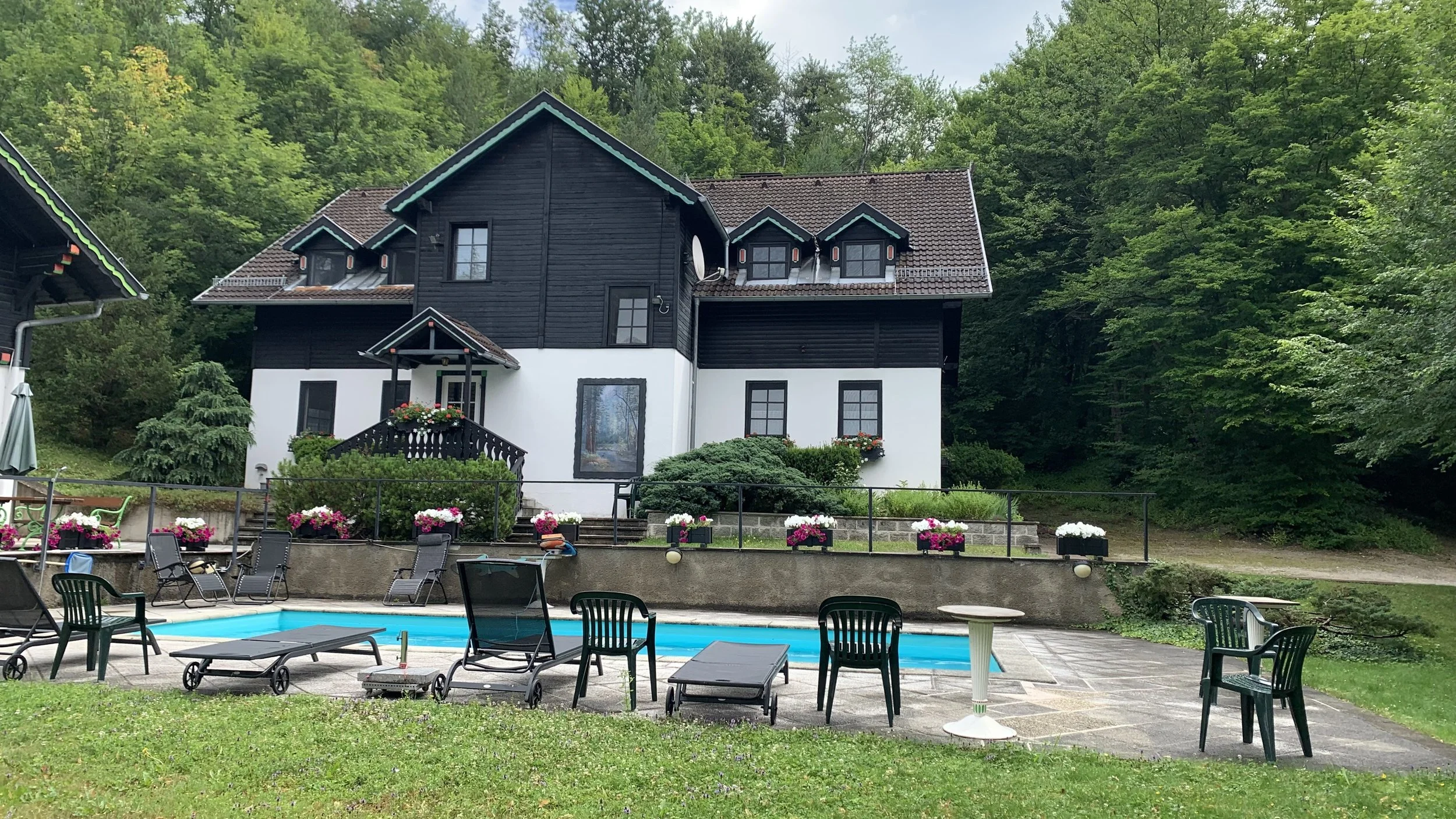 A house with black and white exterior, surrounded by lush green trees and a garden with colorful flowers. In front of the house is a swimming pool area with lounge chairs, tables, and chairs on a paved patio.