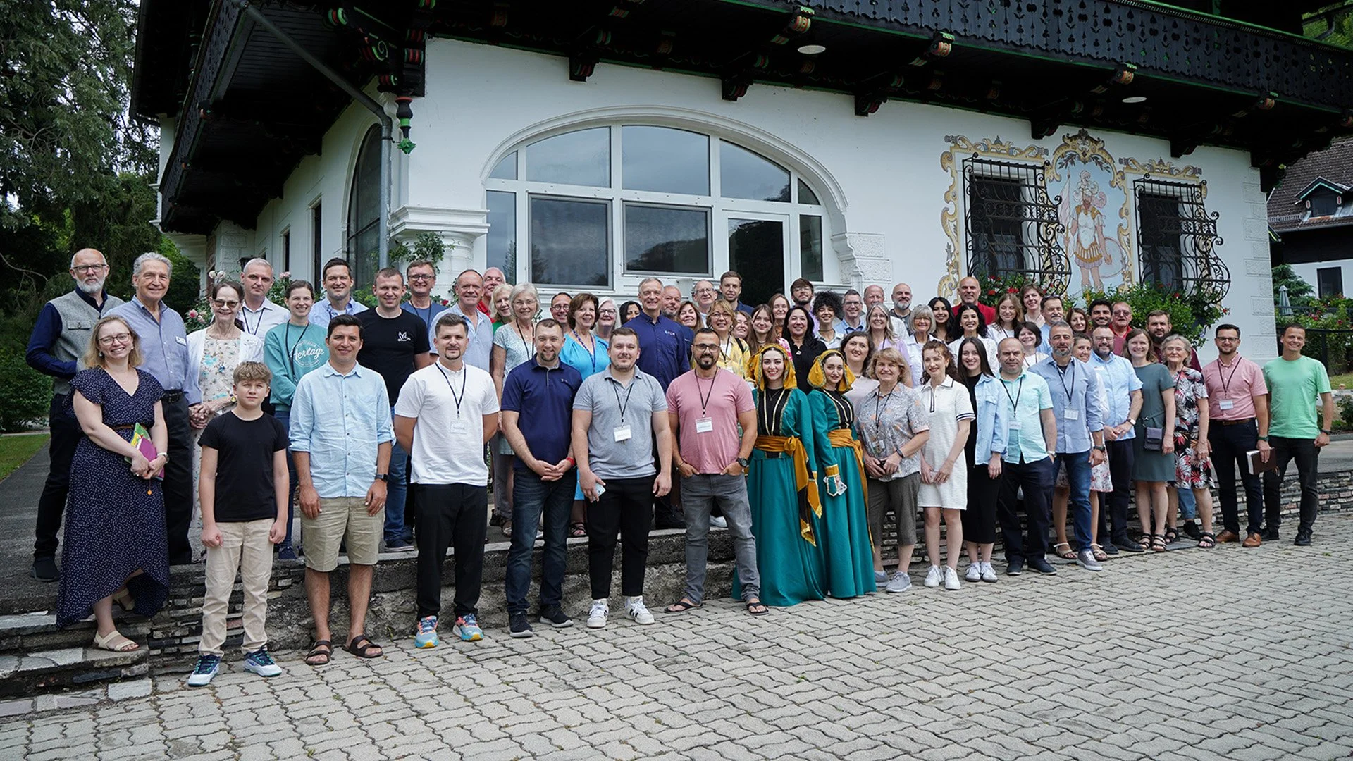 A large group of diverse people standing outdoors in front of a white building with decorative painting and window bars, posing for a group photo.