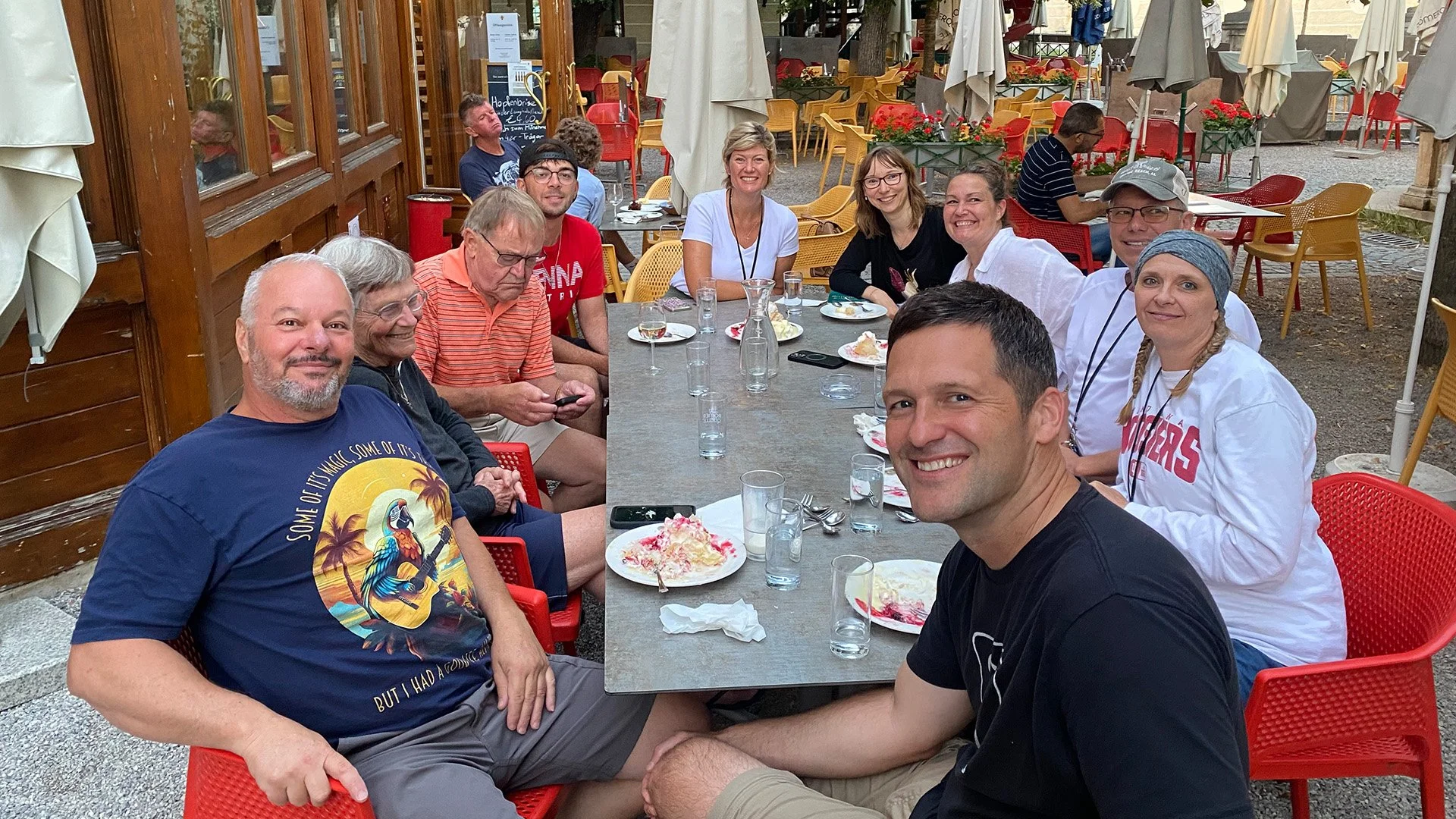 A group of eleven people sitting around a long outdoor dining table at a restaurant, smiling and enjoying dessert with ice cream and drinks, with empty tables and umbrellas in the background.