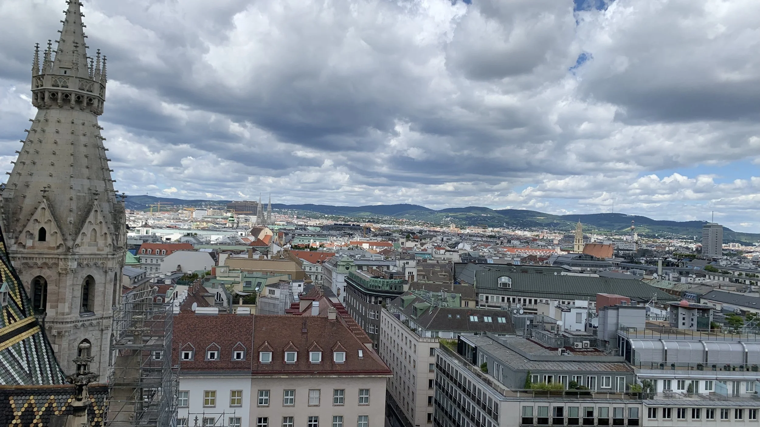 Cityscape view with historic European buildings, church steeples, and rolling hills under a cloudy sky in the background.