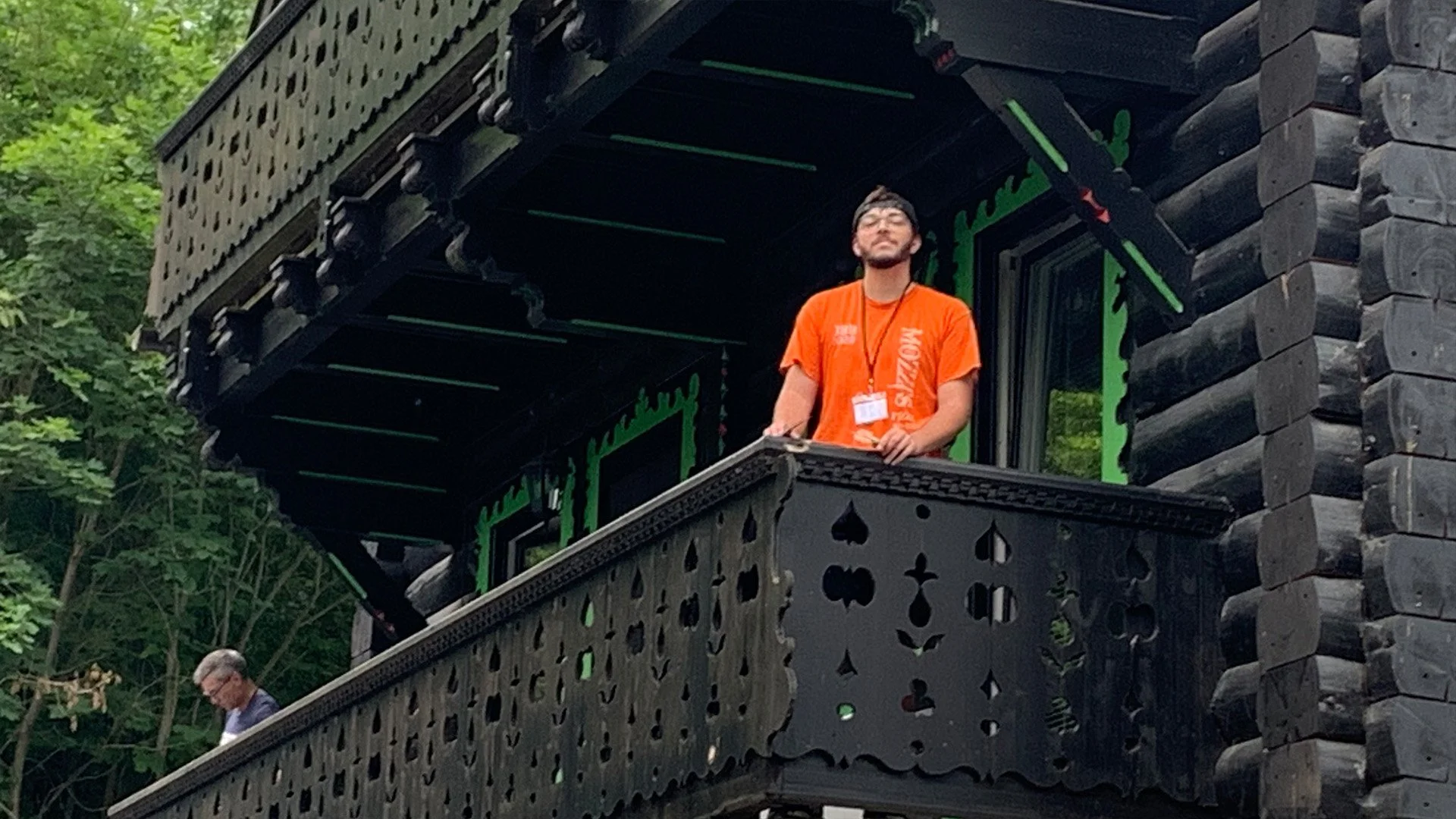 A man with glasses, a beard, and a black cap stands on a wooden balcony of a dark-colored timber house, holding a device in his hand, wearing an orange T-shirt, with trees visible in the background.