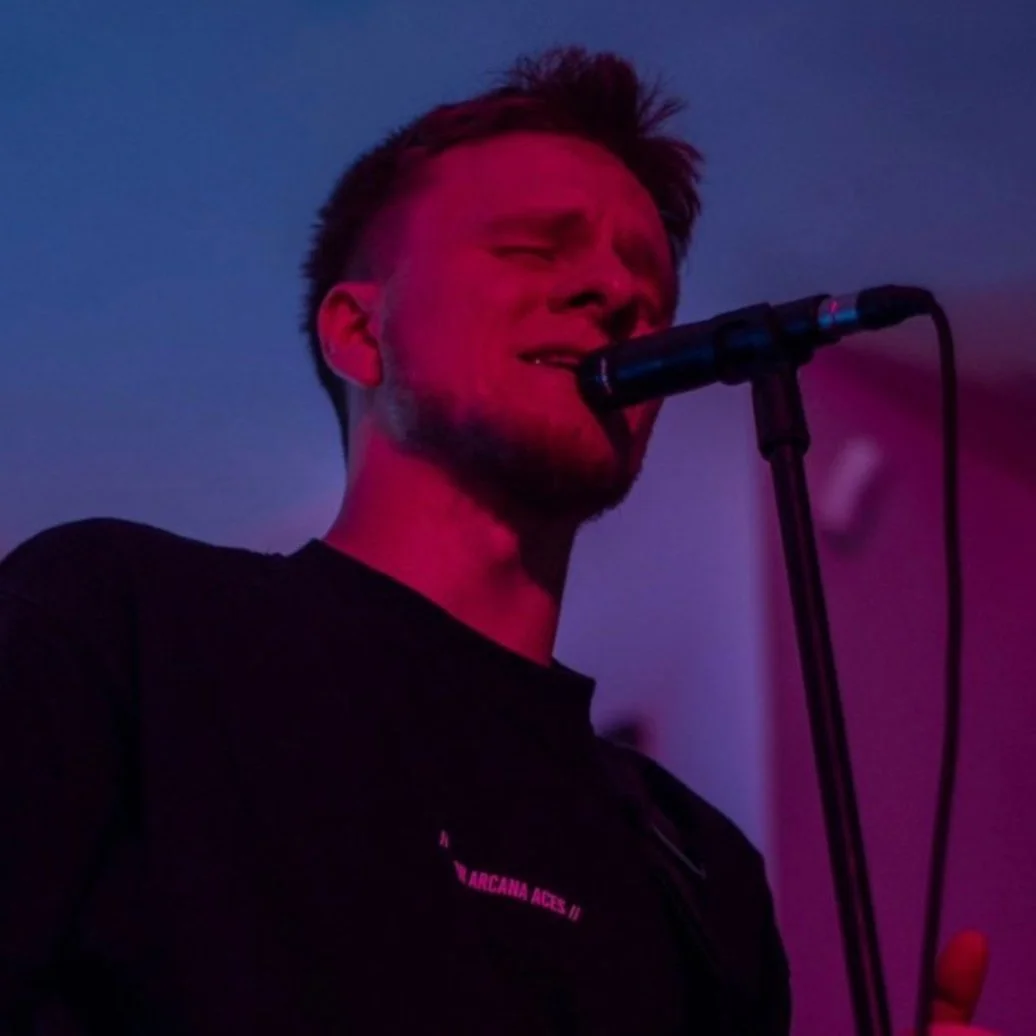 A singer performing passionately with eyes closed, holding a microphone, in a dimly lit room with purple and pink lighting.
