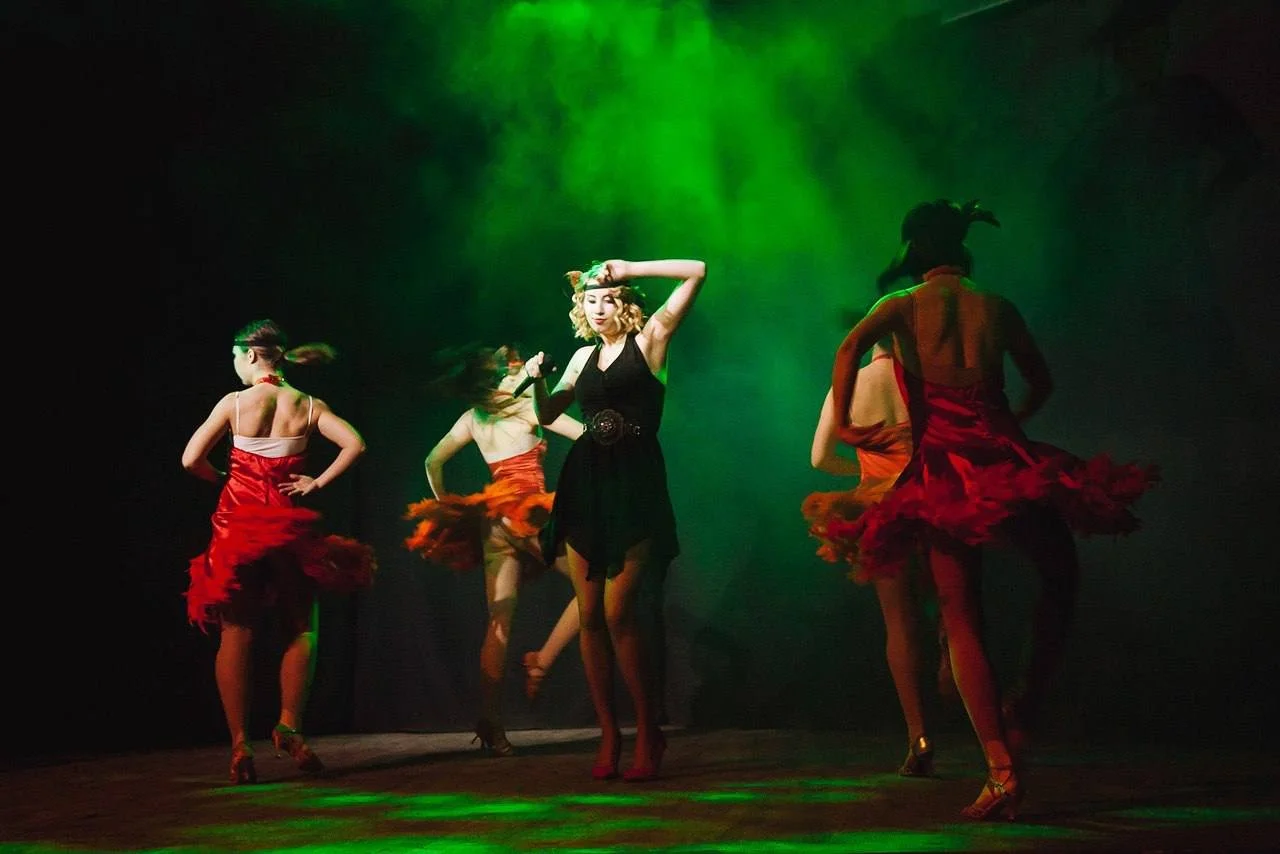 Group of women dancing on stage under green and yellow stage lights, dressed in colorful costumes with red, orange, and black skirts.