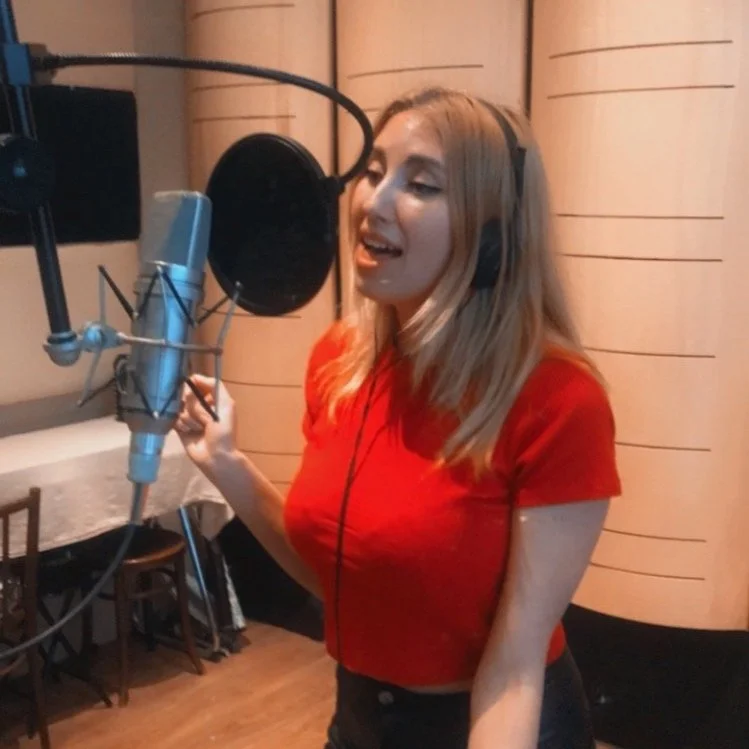 A woman with long blonde hair singing into a professional microphone in a recording studio.